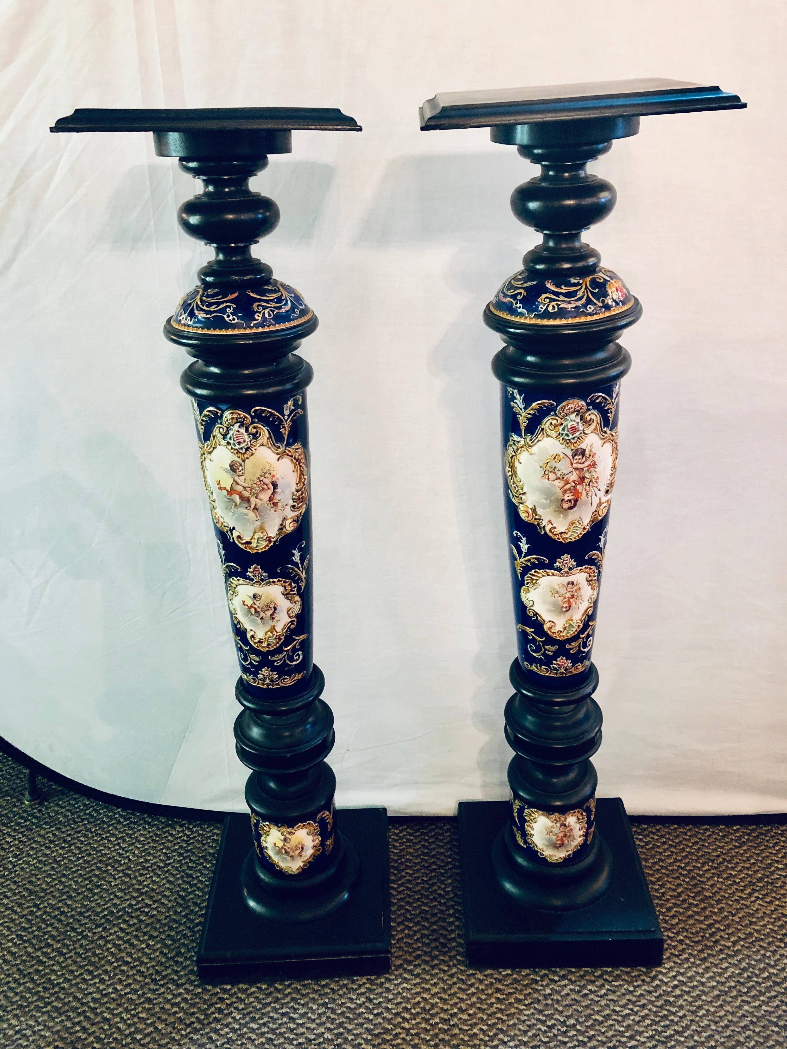 A pair of royal Vienna style porcelain and ebony column pedestals. A fine and highly decorative pair of pedestals each having ebony square table tops on wonderfully hand-painted porcelain cherub scene cobalt blue columns supported by ebony bottoms.