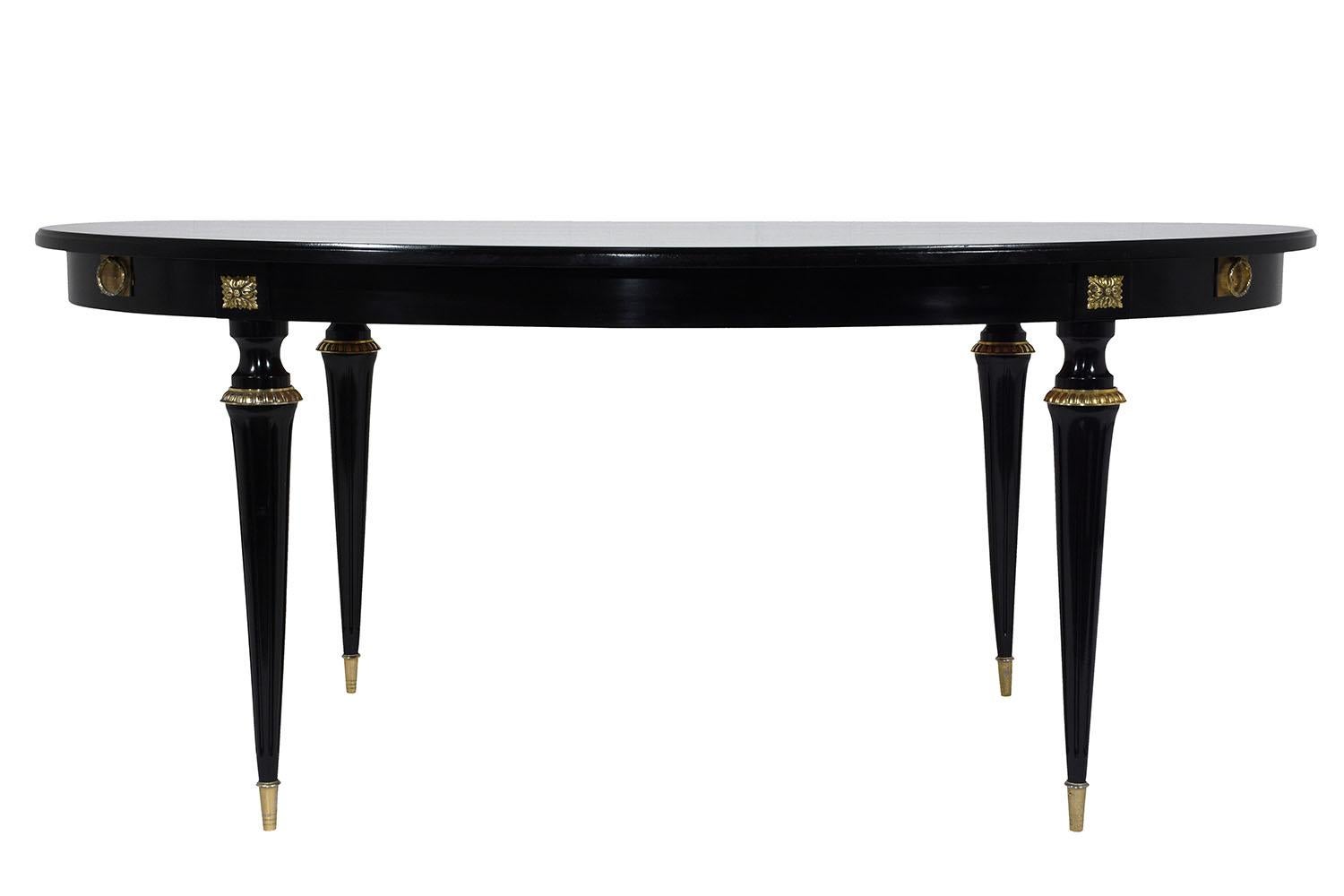 This 1950s French Louis XVI-style dining table is made of mahogany wood ebonized in a rich black color with a lacquered finish. The oval shaped table is elegantly adorned with brass accents along the edge. The carved legs feature fluted details and