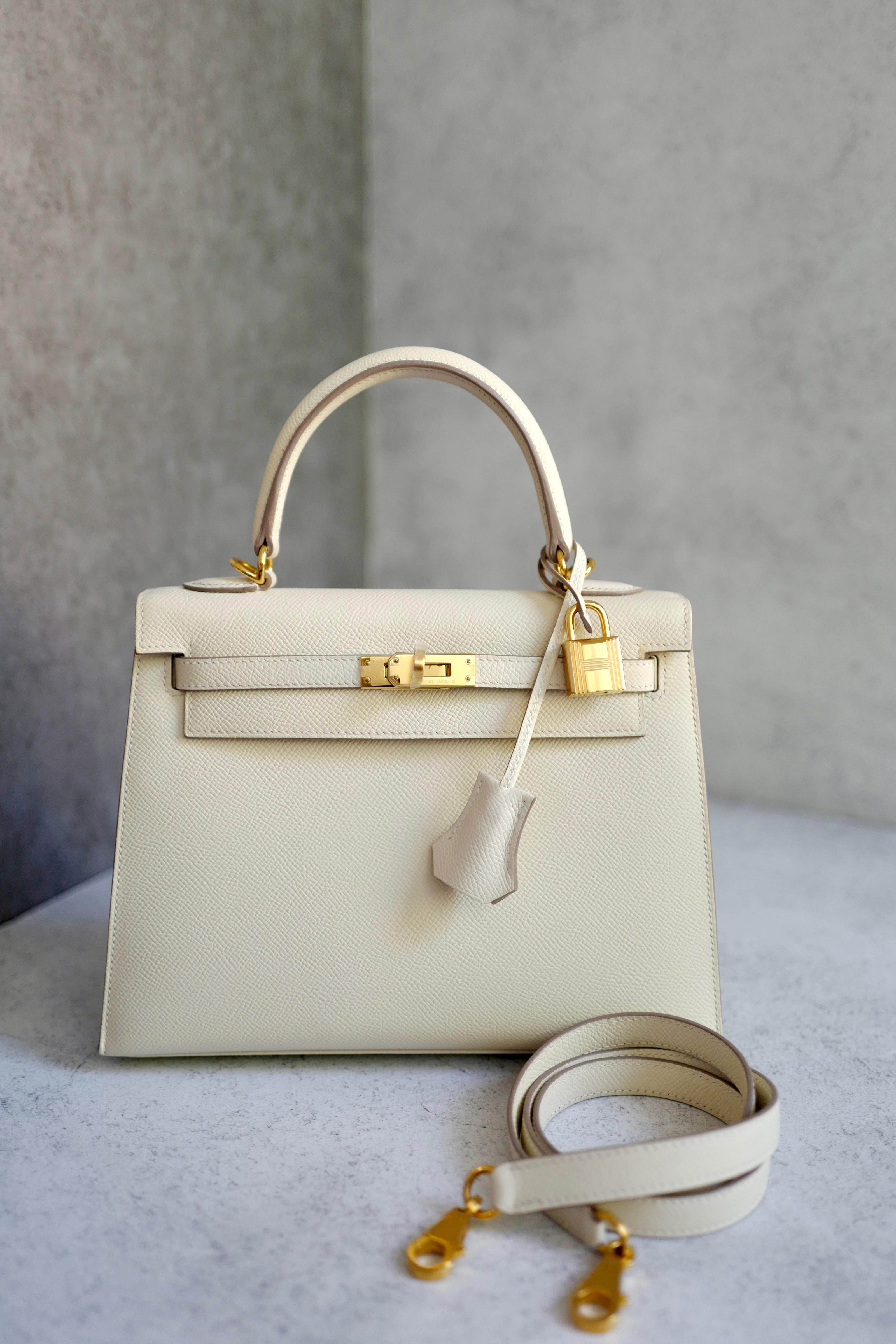 La Kelly 25 Sellier di Hermès in pelle Craie Epsom con finiture dorate è un pezzo elegante e senza tempo della collezione più iconica della Maison. La struttura Sellier conferisce alla borsa una silhouette strutturata dalle linee pulite, mentre la