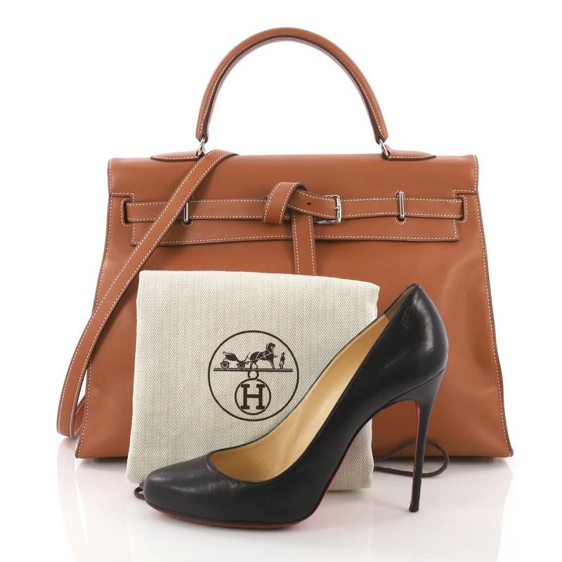 This Hermes Kelly Flat Handbag Gold Swift with Palladium Hardware 35, crafted in gold brown swift leather, features single rolled top handle, unique Hermes etriviere belt strap closure and palladium-tone hardware. Its flap opens to a brown leather