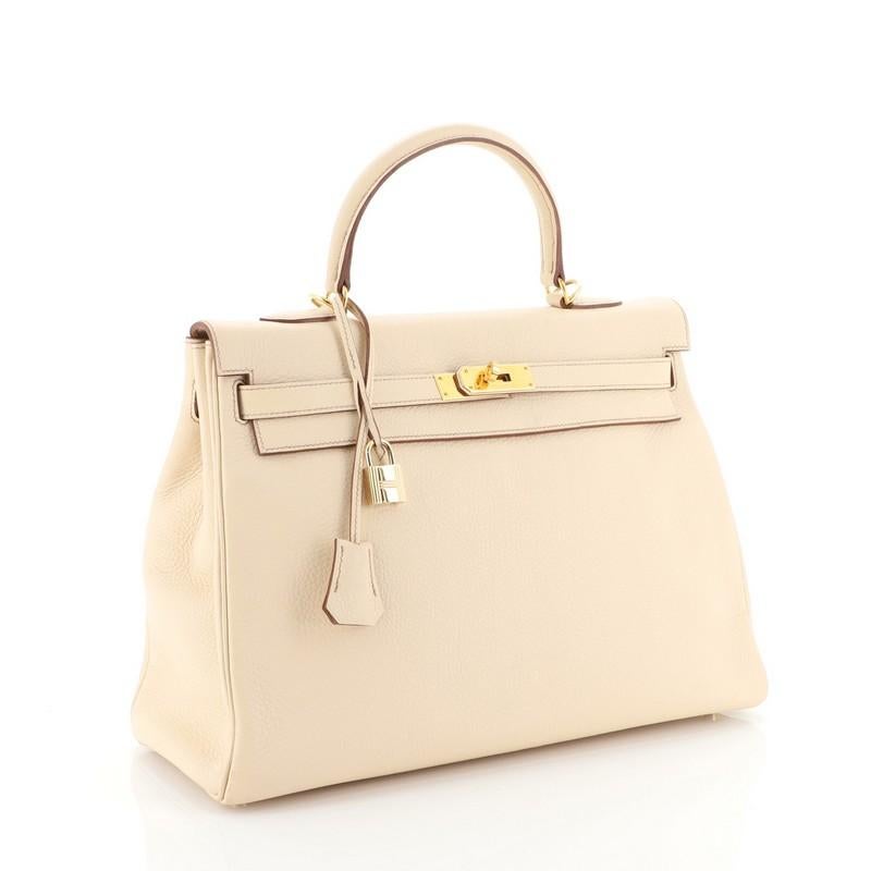 This Hermes Kelly Handbag Beige Clemence with Gold Hardware 35, crafted in Beige neutral Clemence leather, features a single rolled top handle, protective base studs, and gold hardware. Its turn-lock closure opens to a Beige neutral Chevre leather