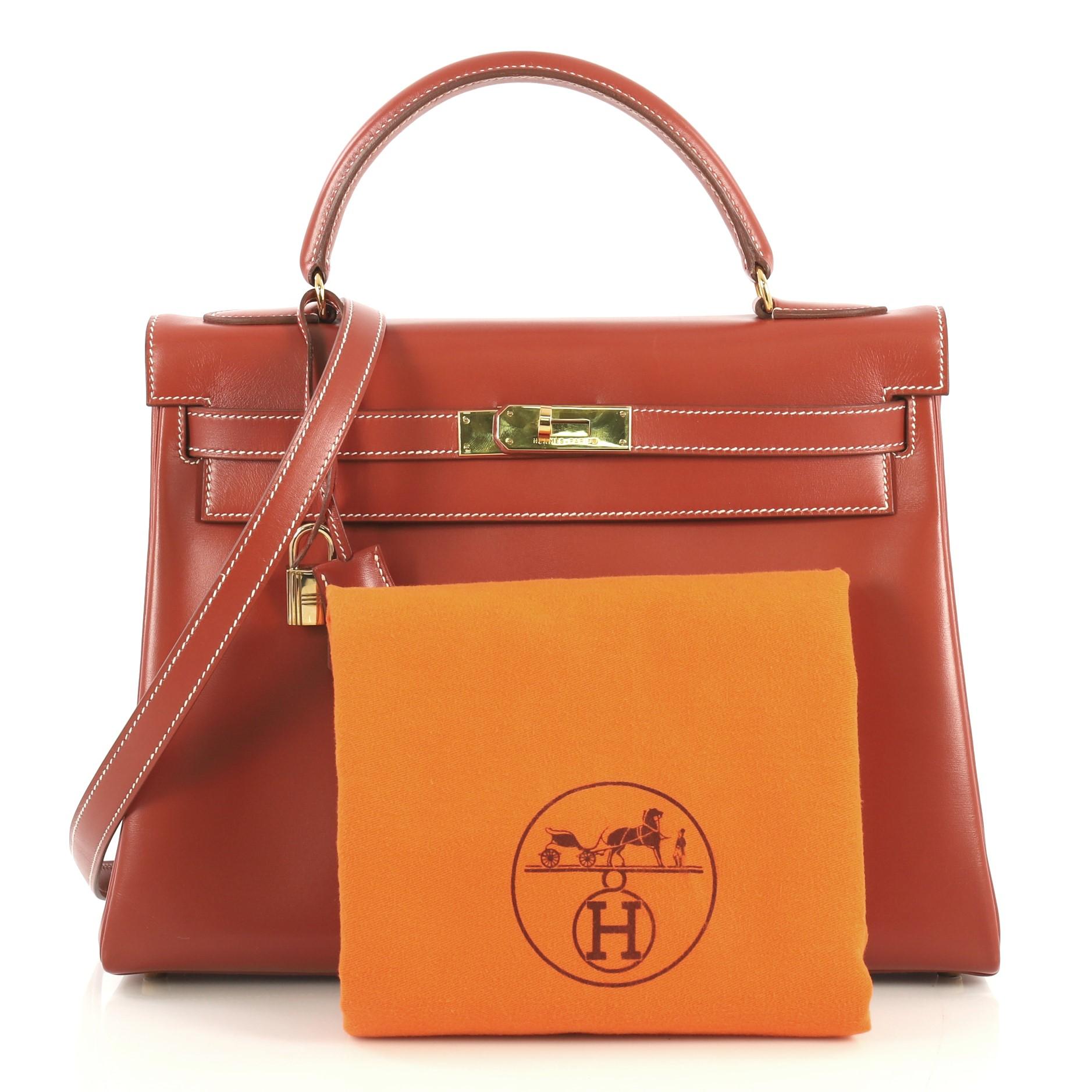 This Hermes Kelly Handbag Brique Box Calf with Gold Hardware 32, crafted in Brique red Box Calf leather, features a single rolled top handle, frontal flap, and gold hardware. Its turn-lock closure opens to a Brique red Chevre leather interior with