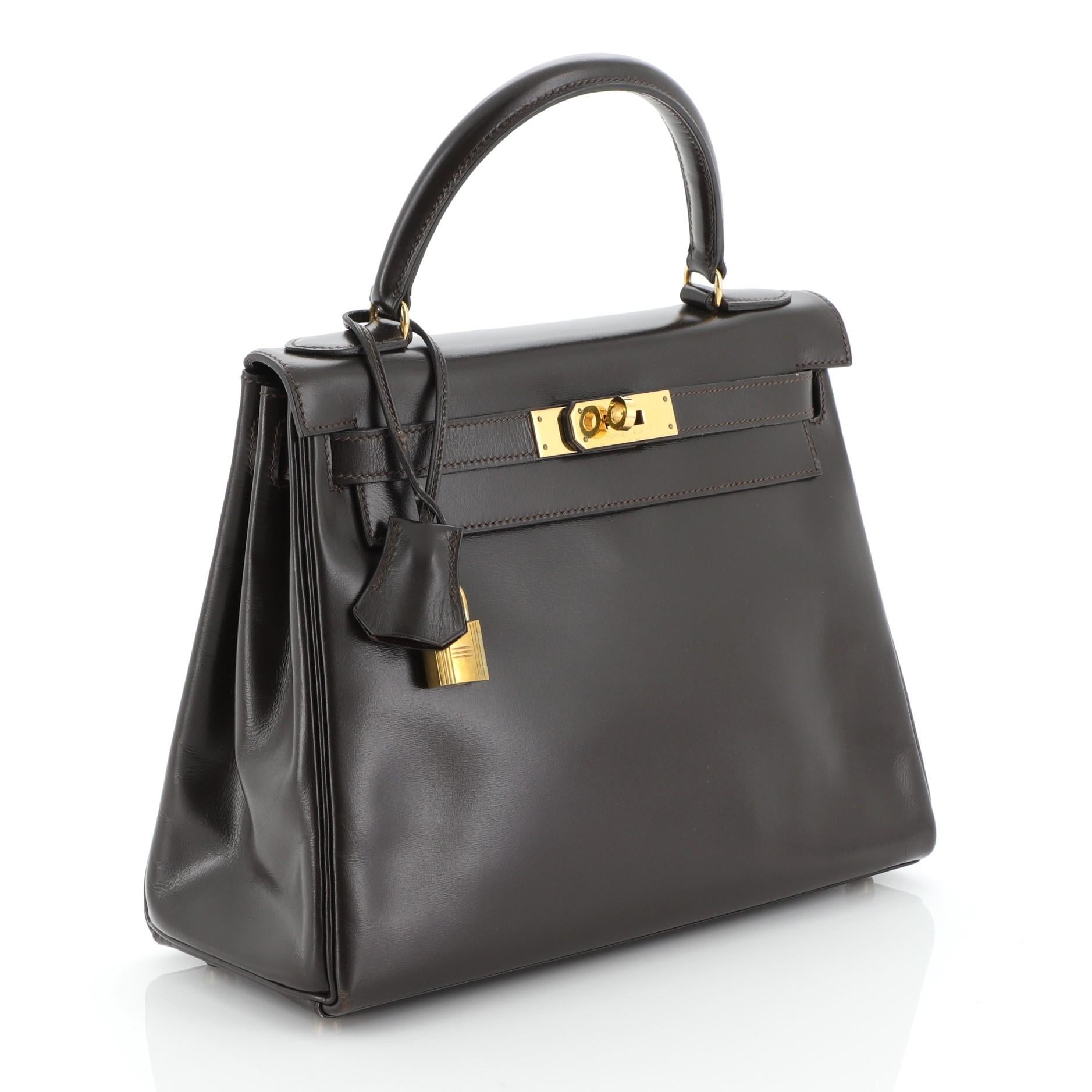 This Hermes Kelly Handbag Ebene Box Calf with Gold Hardware 28, crafted in Ebene brown Box Calf leather, features a single top handle, frontal flap, and gold hardware. Its turn-lock closure opens to an Ebene brown Chevre leather interior with zip
