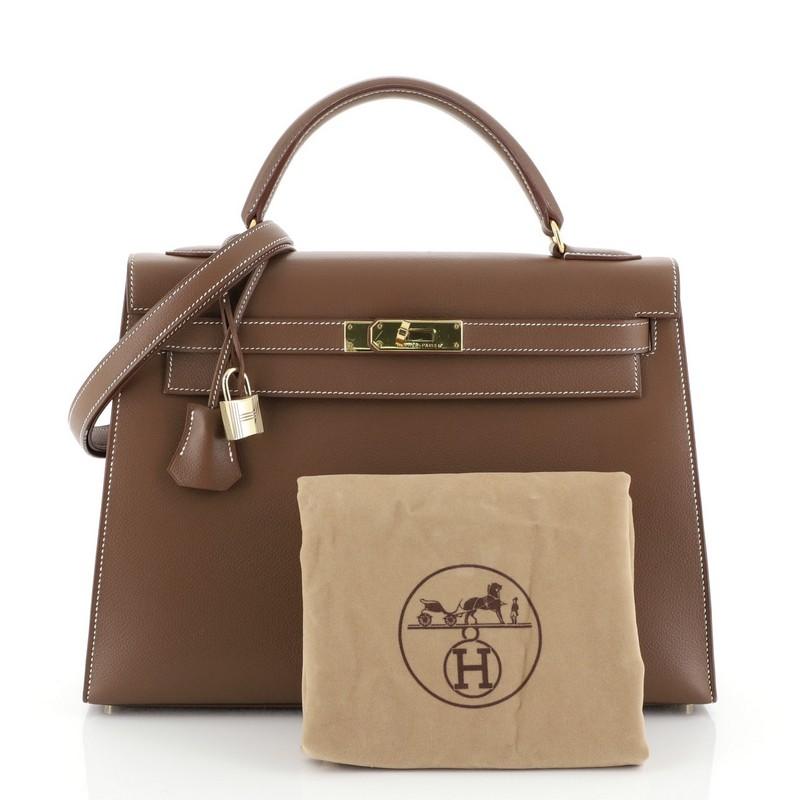This Hermes Kelly Handbag Gold Evergrain with Gold Hardware 32, crafted in Gold brown Evergrain leather, features a single rolled top handle, frontal flap, and gold hardware. Its turn-lock closure opens to a Gold brown Chevre leather interior with