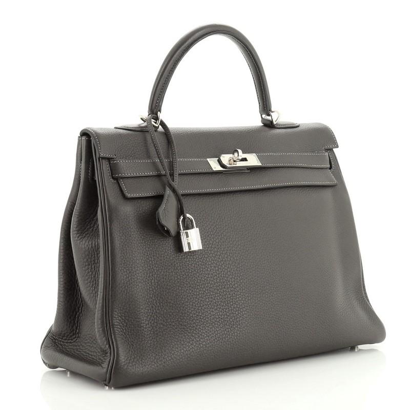 This Hermes Kelly Handbag Graphite Clemence with Palladium Hardware 35, crafted in Graphite grey Clemence leather, features a single rolled top handle, frontal flap, and palladium hardware. Its turn-lock closure opens to a Graphite grey Chevre