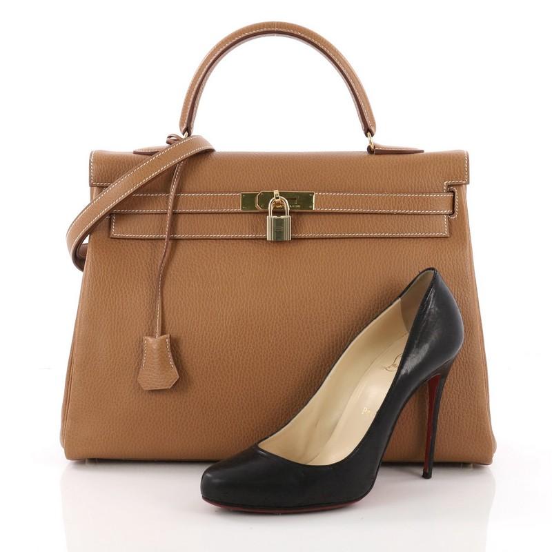 This Hermes Kelly Handbag Natural Ardennes with Gold Hardware 35, crafted from natural brown Ardennes leather, features a single rolled top handle, frontal flap, protective base studs, turn lock closure and gold-tone hardware. It opens to a brown