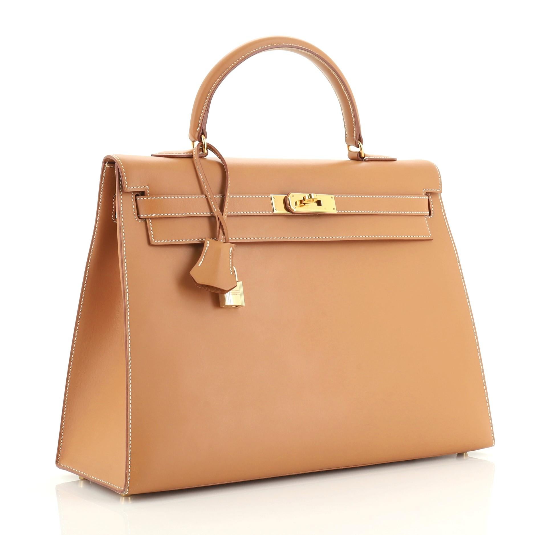 This Hermes Kelly Handbag Natural Chamonix with Gold Hardware 35, crafted from Natural brown Chamonix leather, features a single top handle, frontal flap, protective base studs, and gold hardware. Its turn-lock closure opens to a Natural brown