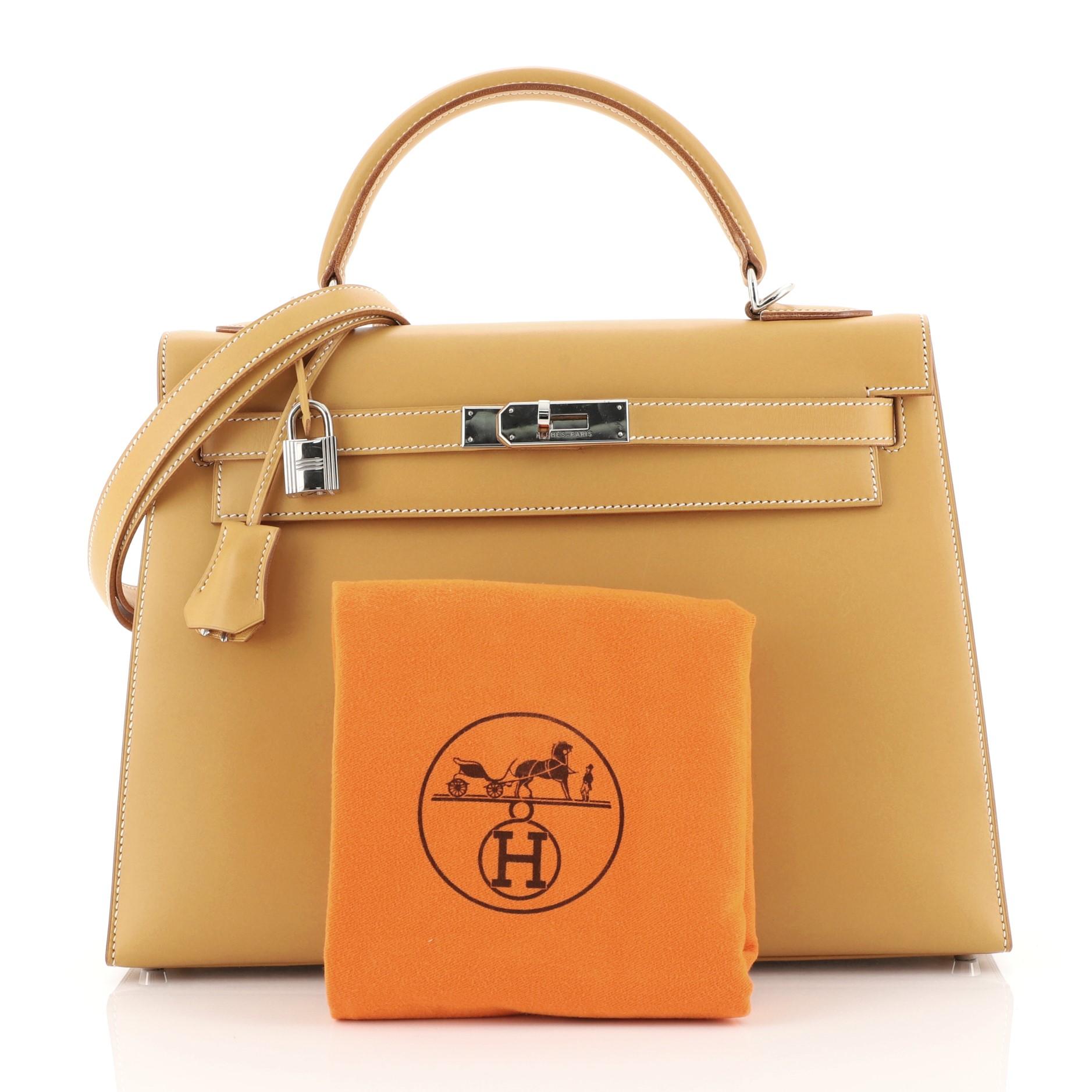 This Hermes Kelly Handbag Natural Chamonix with Palladium Hardware 32, crafted in Natural brown Chamonix leather, features a rolled top handle, protective base studs, and palladium hardware. Its turn-lock closure opens to a Natural brown Chevre