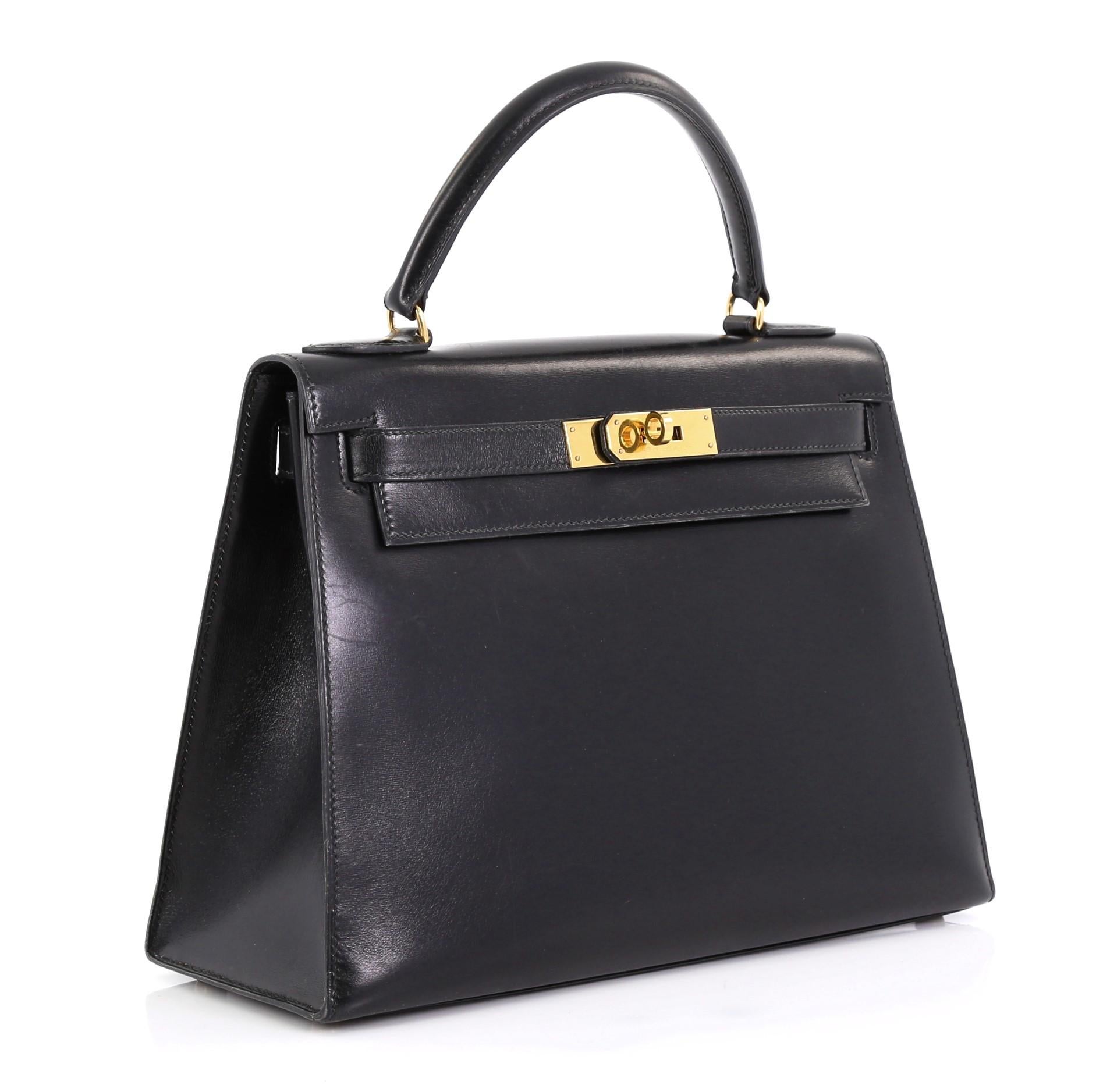 This Hermes Kelly Handbag Noir Box Calf with Gold Hardware 28, crafted in Noir black Box Calf leather, features a single rolled top handle, protective base studs, and gold hardware. Its turn-lock closure opens to a Noir black Chevre leather interior