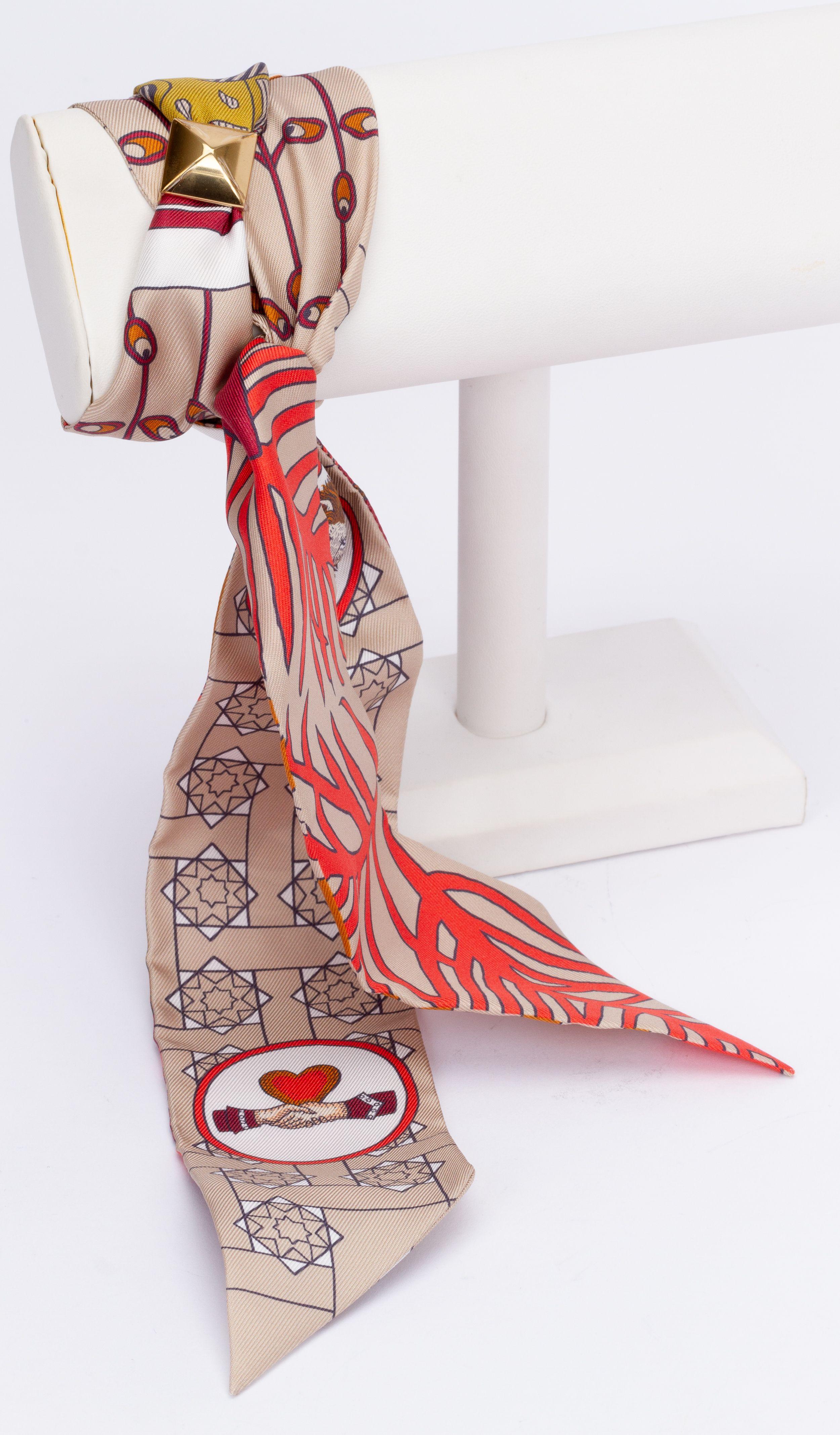 New Hermès La Danse Des Amazones Twilly. The scarf is made out of silk and the main color is beige and coral. The print shows animals like an owl. The piece comes with a scarf ring, original box and ribbon.