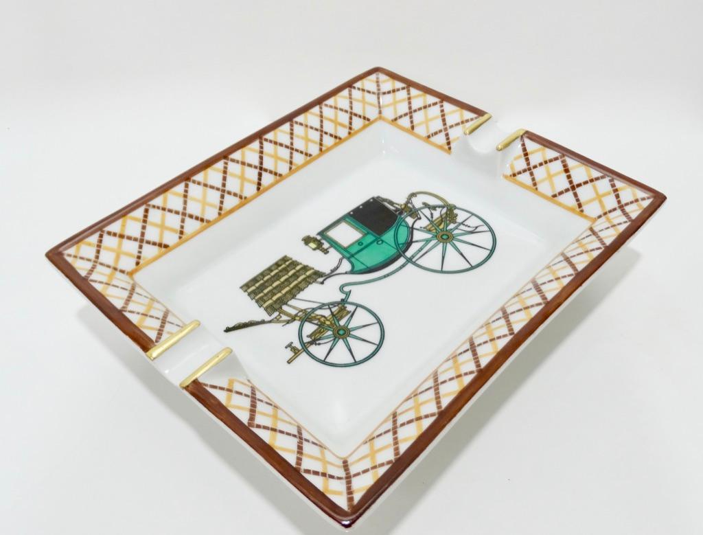 Decorate in style with this amazing Hermés tray! Circa late 20th century, this tray is crafted from porcelain and features an emerald Calèche or carriage motif surrounded by a tan and brown crossed trim with gold metallic accents. Underside is