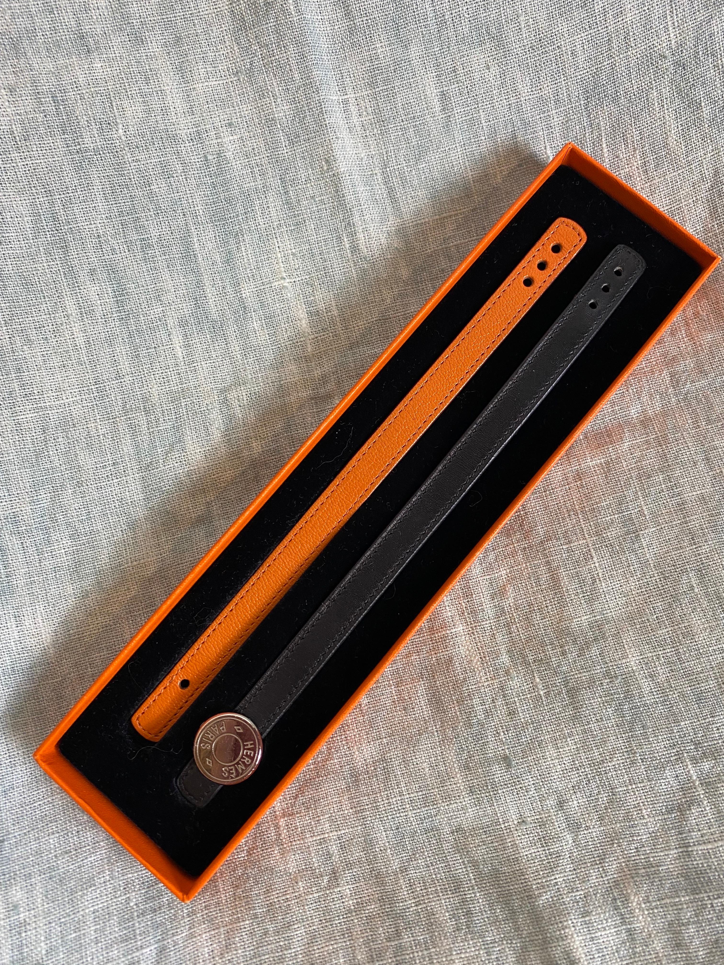 Hermes leather bracelets one black and one orange with silver metal clasp, opens by turning the top part and can be swapped with the two bracelets and used to adjust the length having 3 holes. 
In excellent condition, it shows only a few small signs
