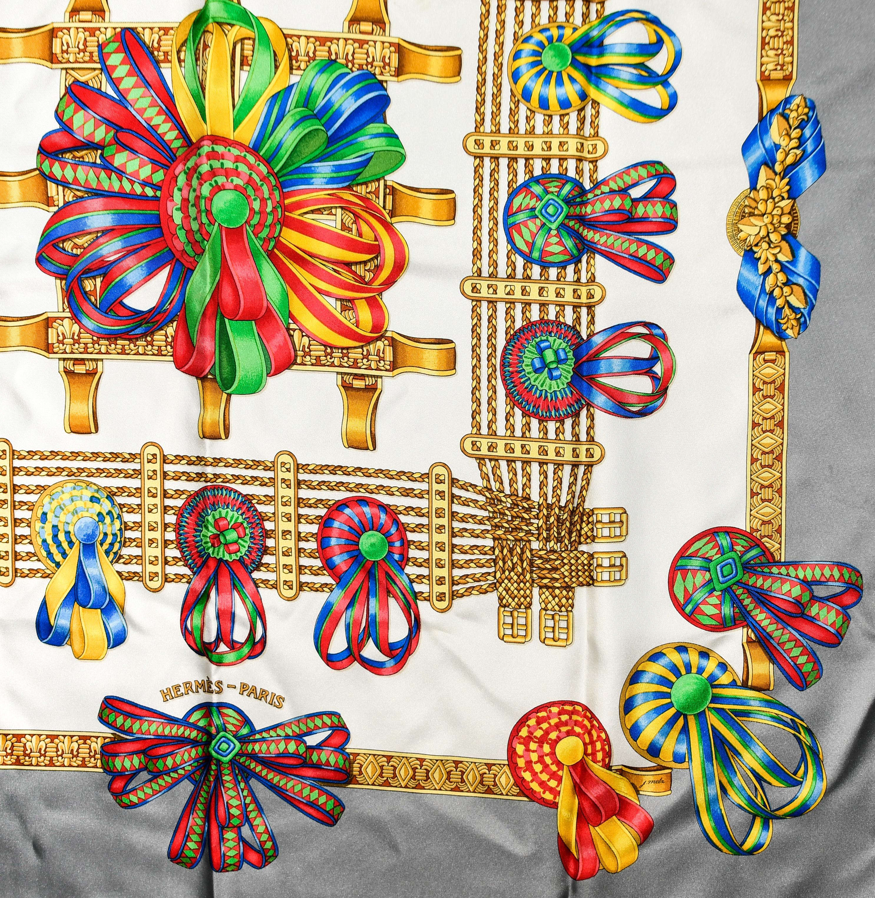 Hermes 100% silk square scarf that features a bright jewel tone graphic design of ribbons on a gold framework, layered over a white background.  These ribbons represent, in the equestrian world, the achievement of the magnificent horses and their