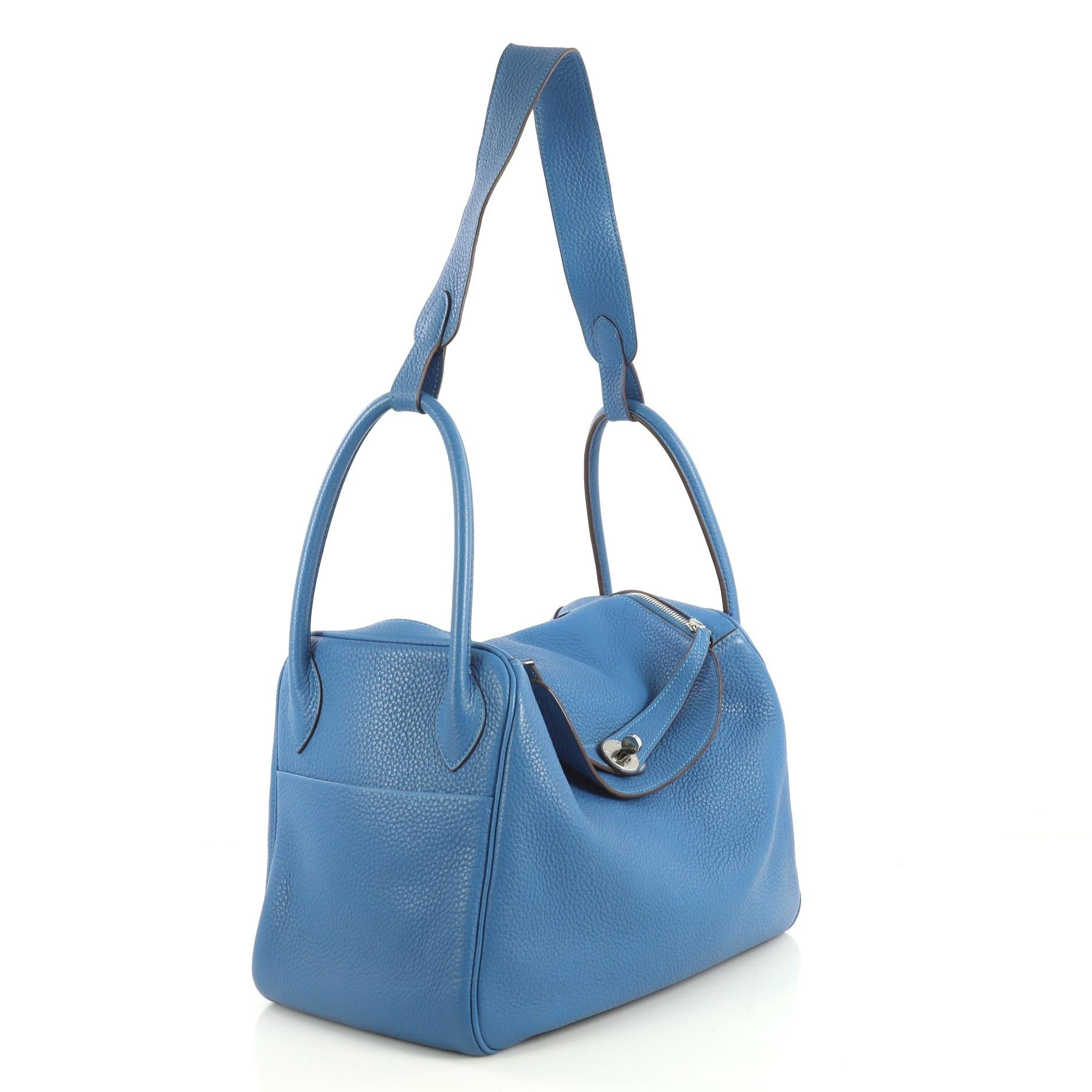 This Hermes Lindy Handbag Clemence 34, crafted from Mykonos blue Clemence leather, features dual rolled handles with a connecting long strap, two side slip pockets, protective base studs, and palladium hardware. Its double zip closure opens to a