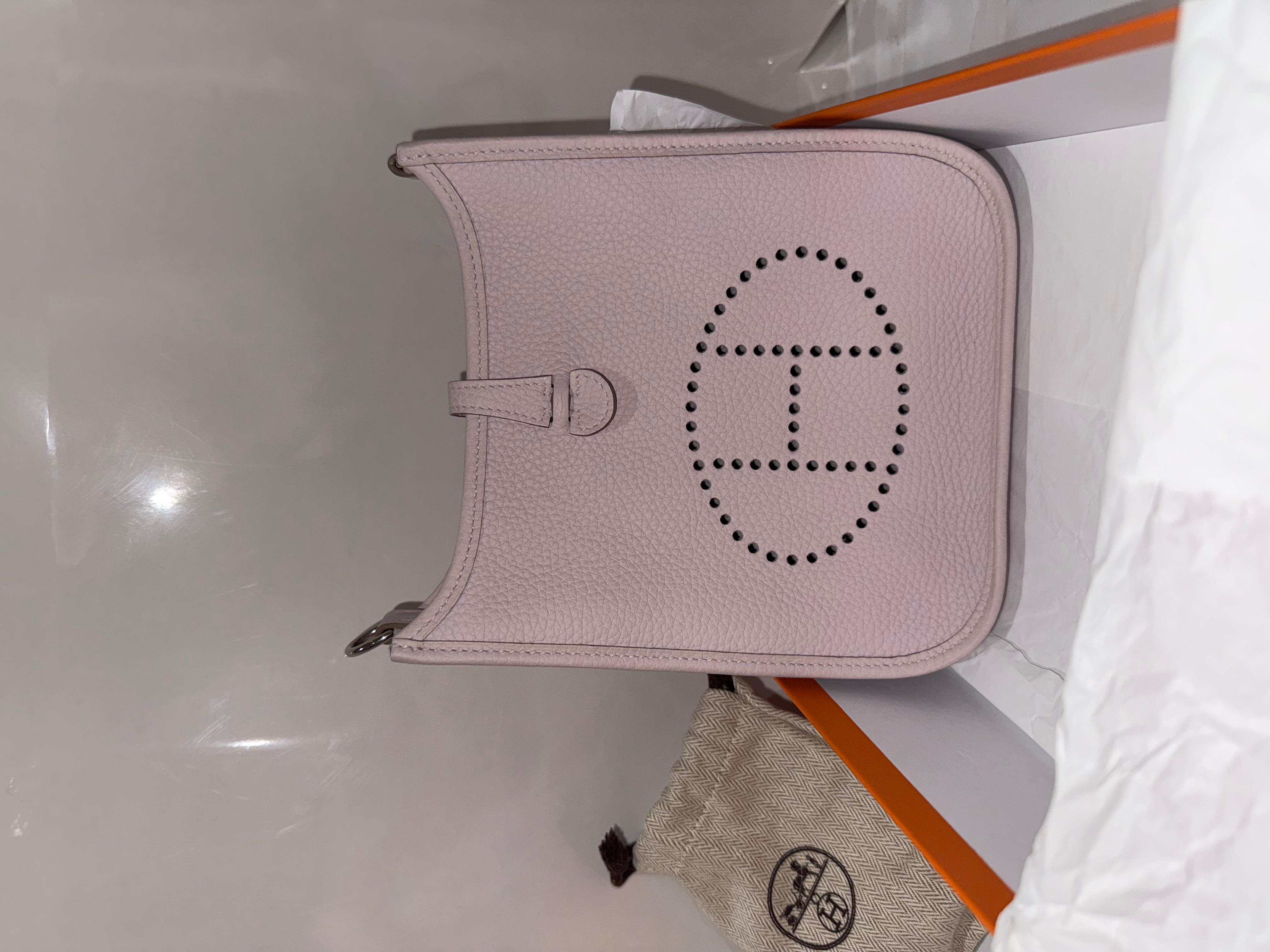Hermes mini Evelyne 16 Taurillon Clemence in color malva e palladio. Questa elegante borsa messenger piccola è realizzata in pelle di vitello Clemence testurizzata in rosa chiaro. Presenta un ovale Hermes H. traforato sul davanti e una tracolla in