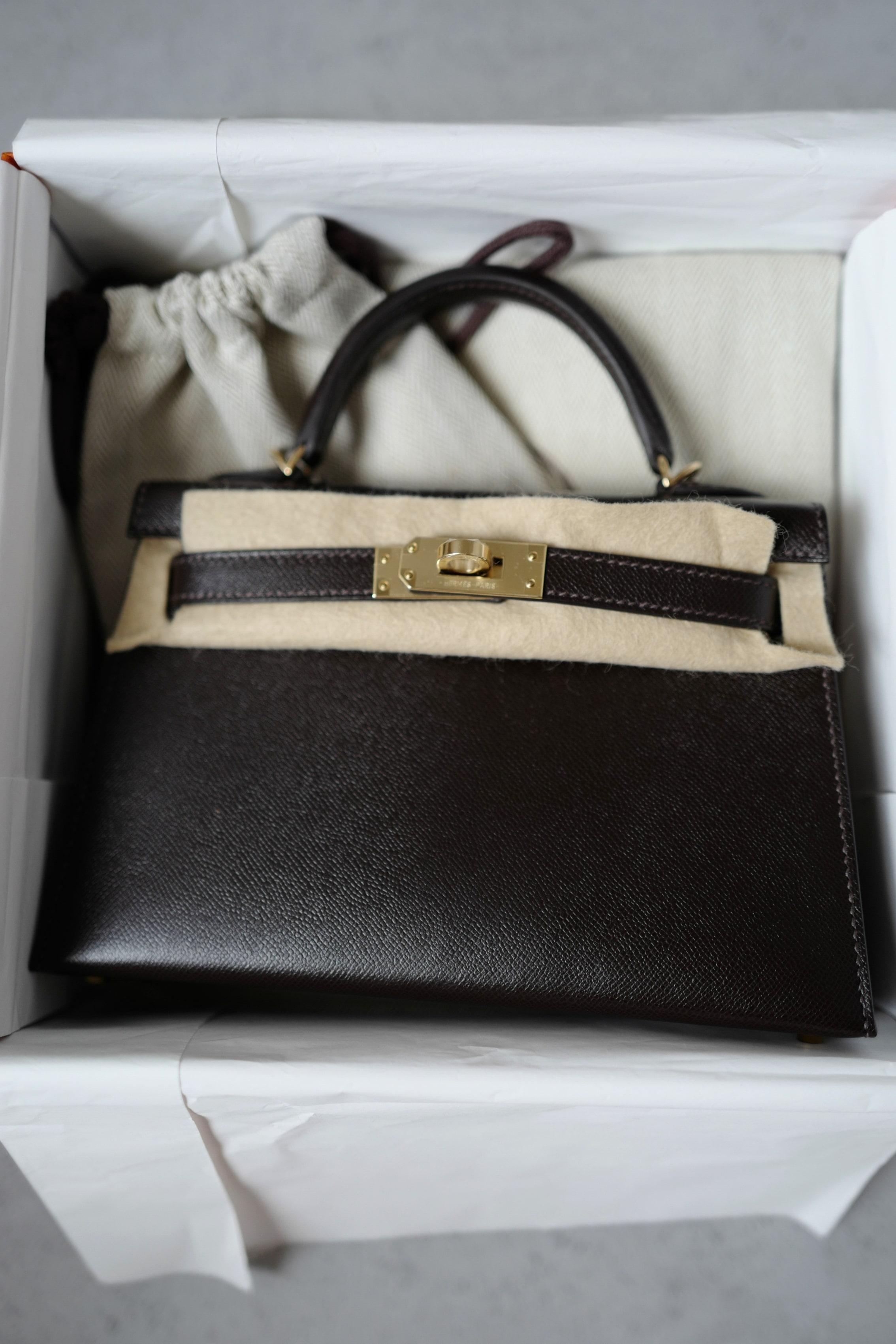 La Mini Kelly 20 di Hermès in pelle Bai Brun Veau Madame è un capolavoro di eleganza raffinata e sofisticatezza senza tempo. Realizzata in pelle di vitello a grana fine, questa squisita borsa presenta una silhouette strutturata con una sottile