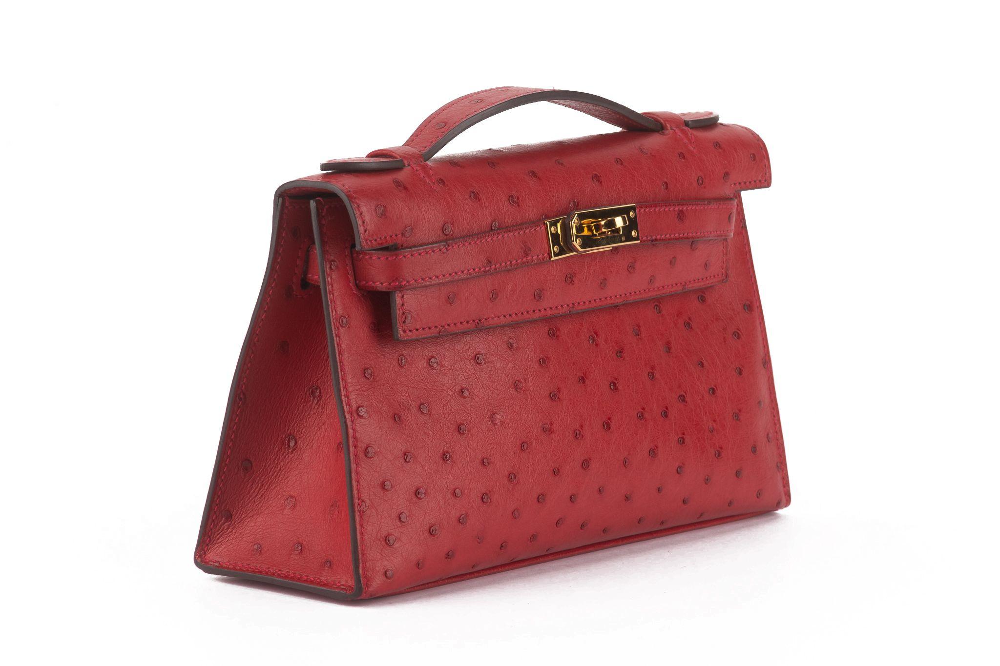 Cette pochette en cuir d'autruche Kelly de couleur rouge présente une teinte rouge vif. Ce sac à main est doté d'une petite poignée supérieure, d'un rabat croisé et d'une fermeture à lanière avec un cadenas doré. Il s'ouvre sur un intérieur en cuir
