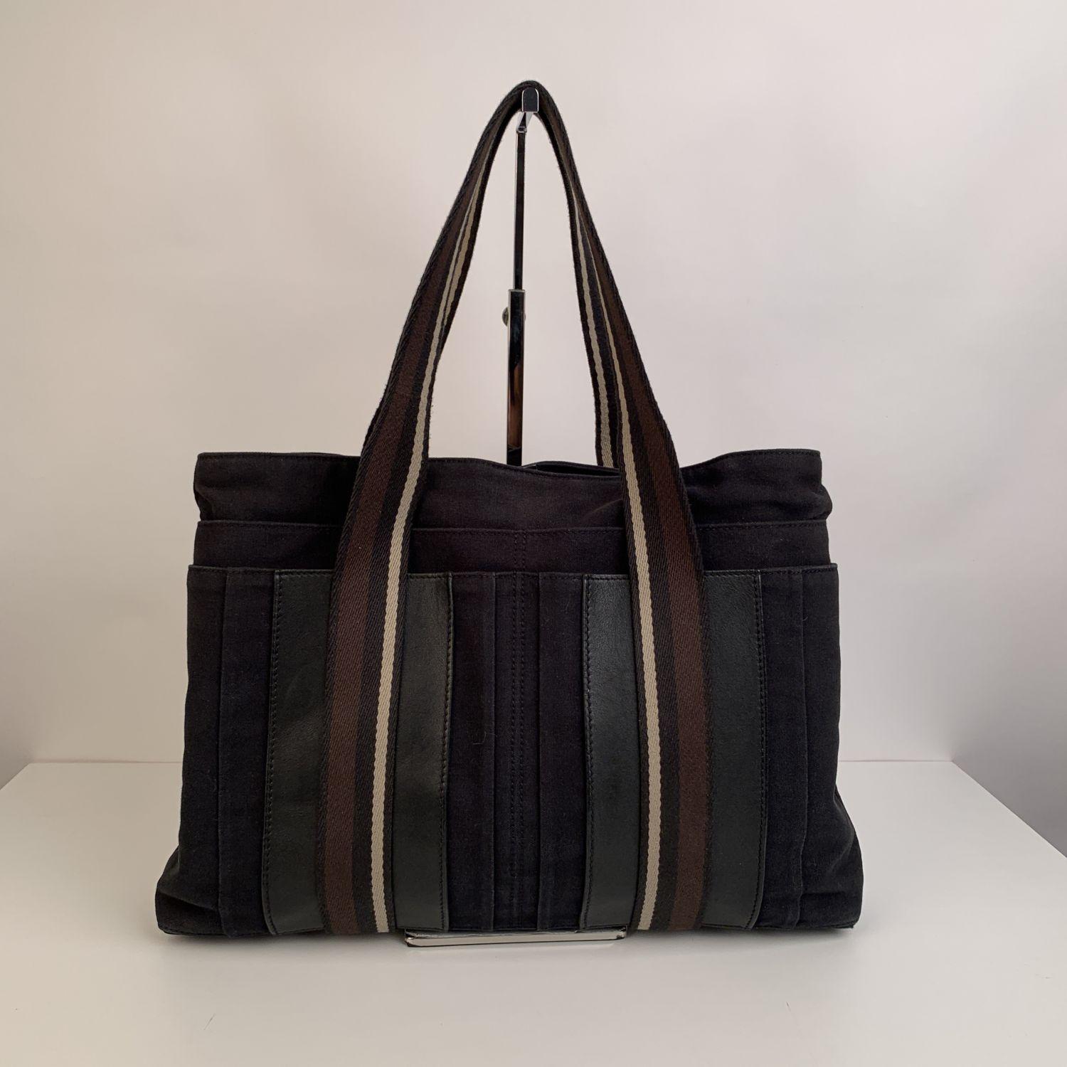 Hermès Horizontal Troca MM bag crafted in a black cotton canvas with genuine leather trim. Brown and white stripes detailing. It features double top handles, 4 front slip pockets and upper button closure. Canvas lining and and 1 zip pocket inside.