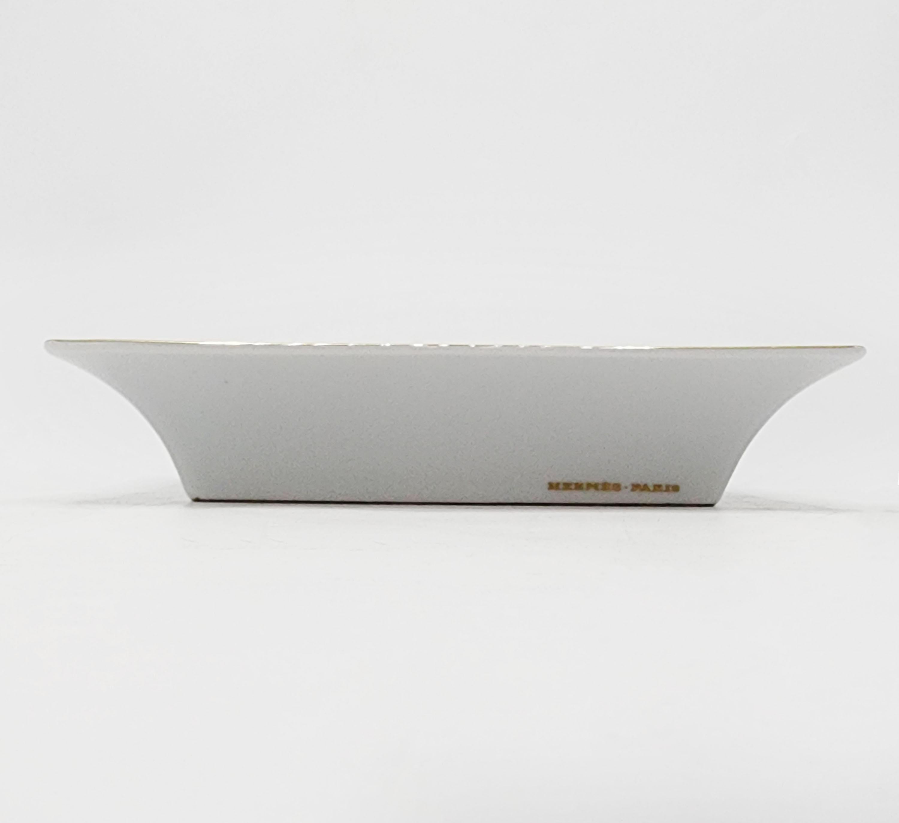 Posacenere in porcellana Hermes Paris
Il posacenere ha un design elegante e classico, con una forma rettangolare e bordi leggermente arrotondati. La sua base è di un grigio tenue, che contrasta armoniosamente con i dettagli dorati che adornano il