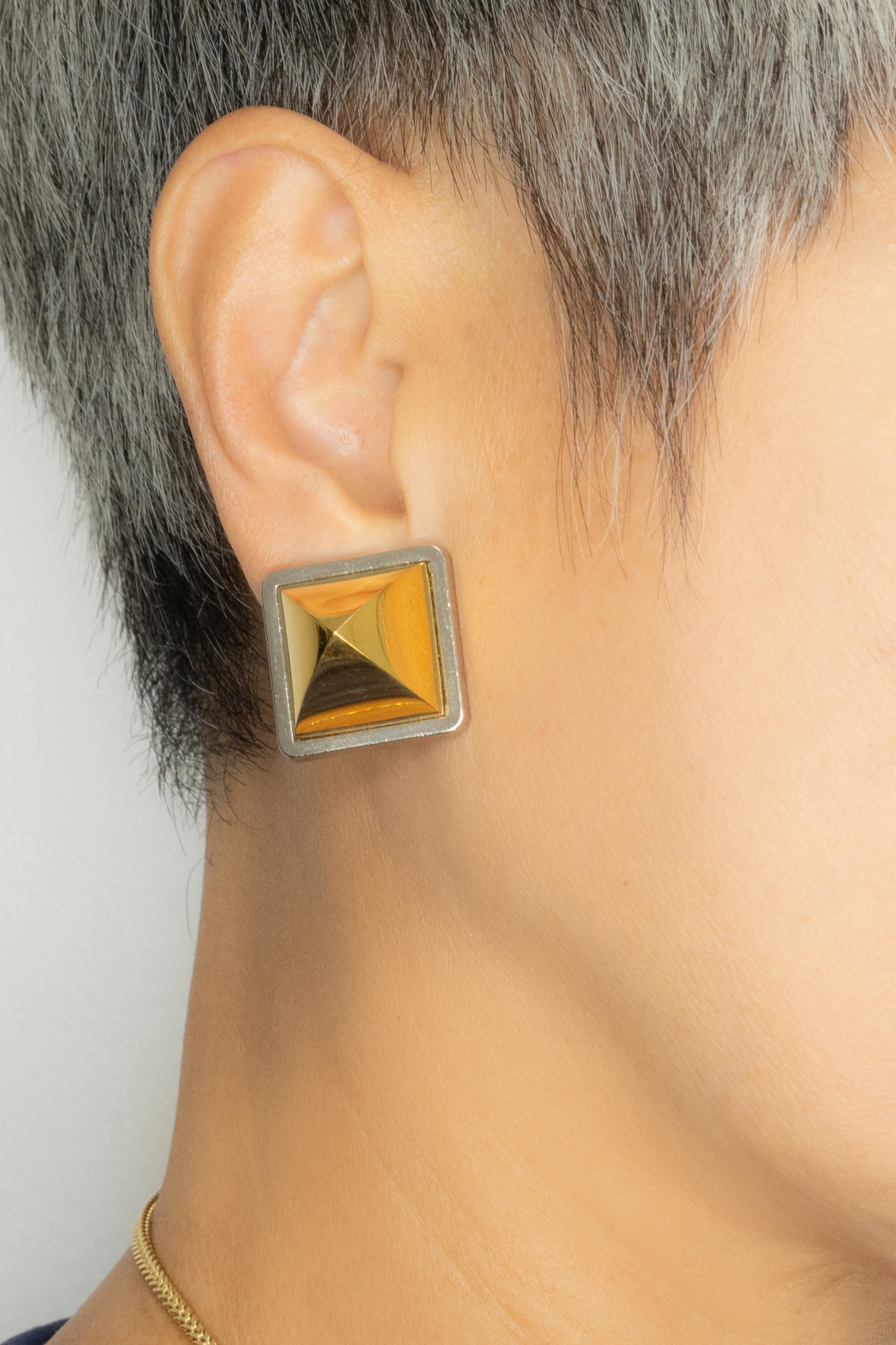 A striking pair of vintage Hermès Paris earrings showcasing the maison’s signature bold, architectural aesthetic. Each earring features a raised, pyramid-style  in gleaming gold-tone framed by a sleek silver-tone border, creating a chic contrast of