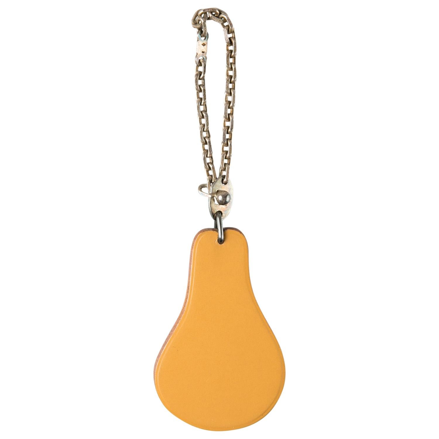 Introduce a fun style to your classic totes with this amazing bag charm from Hermes. Crafted from tan-colored leather into a pear shape, the charm is adorned with a green leaf motif for an ideal replication along with a silver chain attachment and a