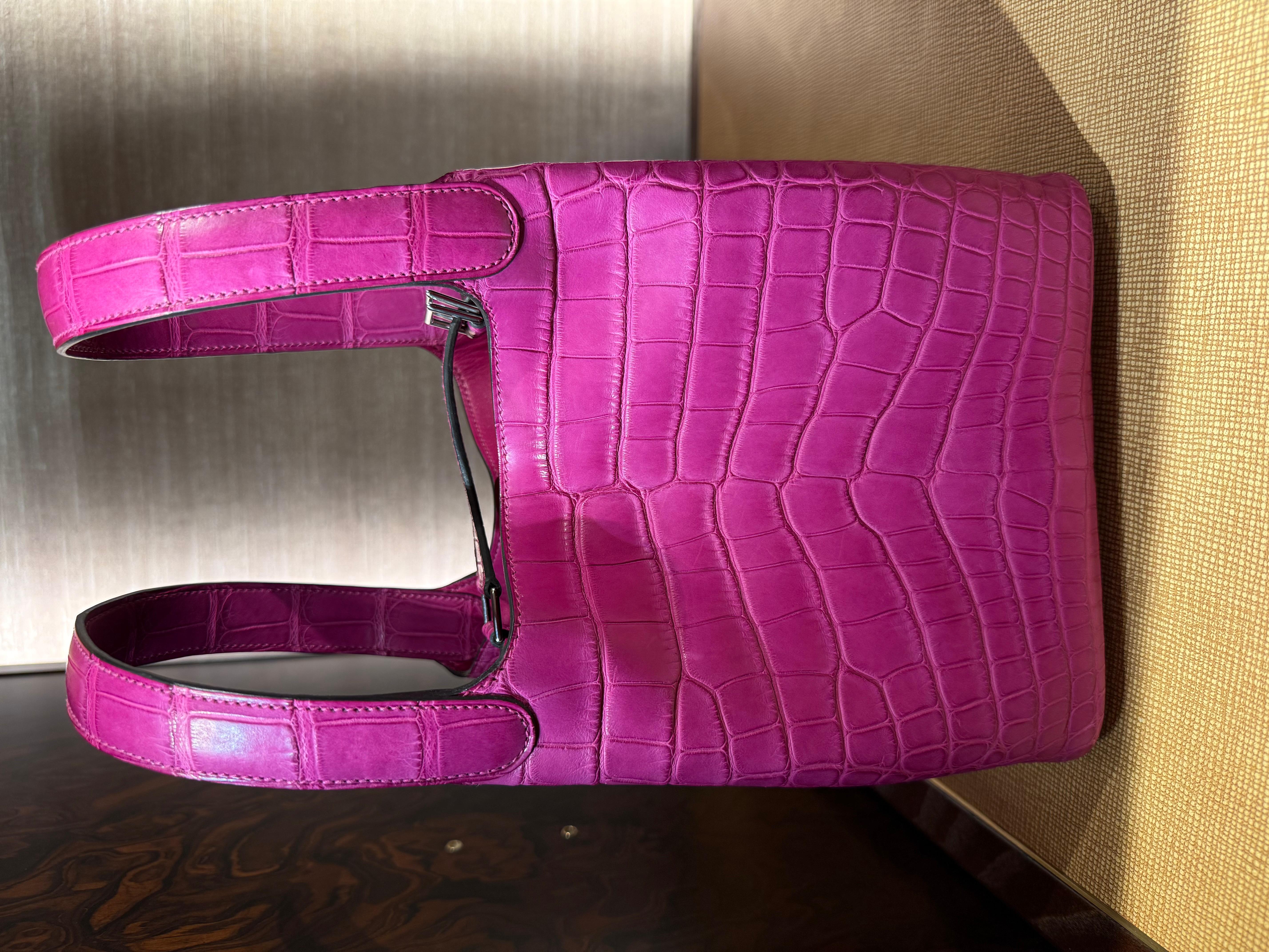 Le Picotin 18 d'Hermès est en cuir Mississippiensis en alligator mat Scheherazade rose, avec des accessoires en palladium, des surpiqûres tonales et deux anses supérieures. Timbre W, production 2024.

L'intérieur est doublé de cuir de martinet rose,