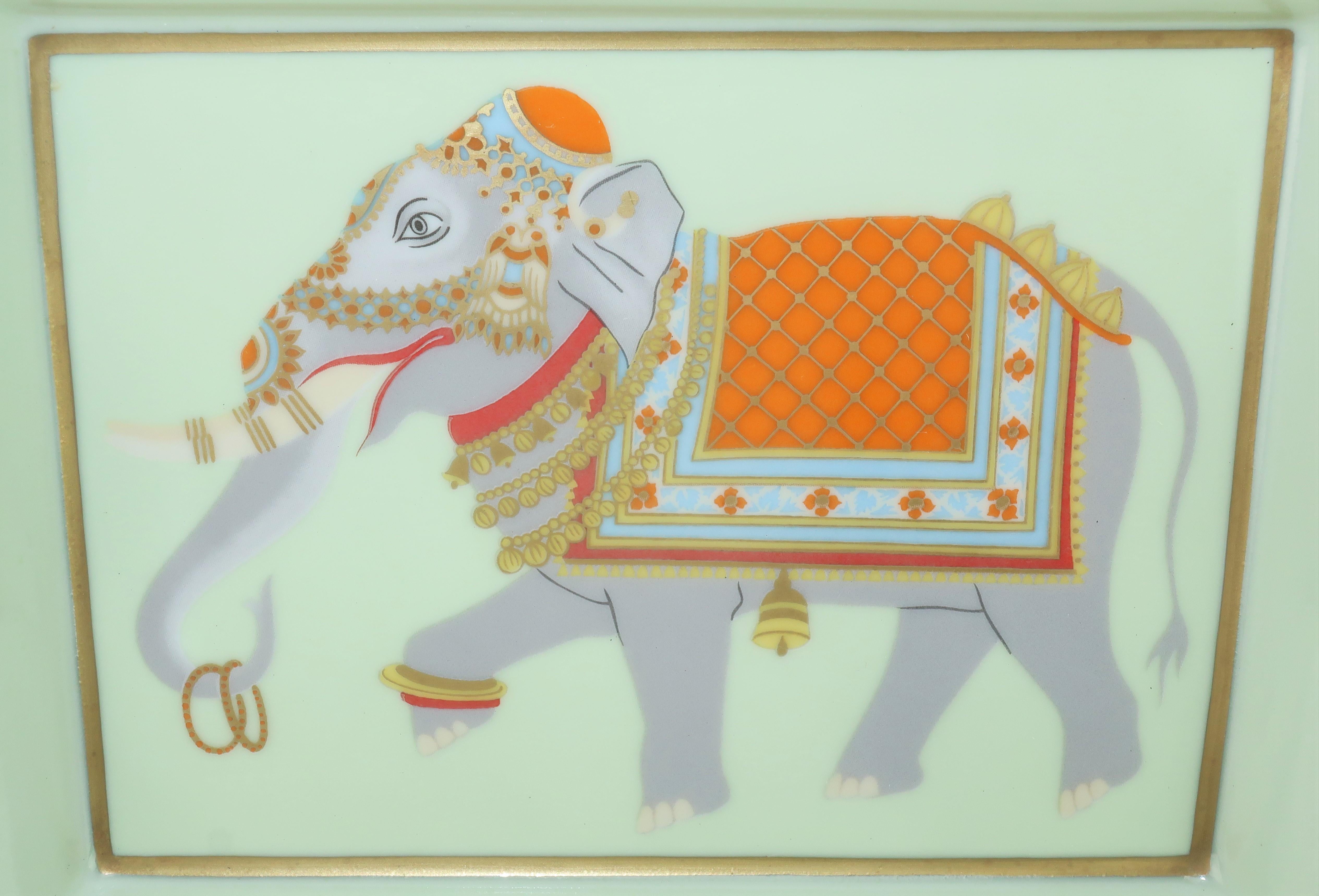 A beautifully exotic Hermès porcelain ashtray in shades of orange, pale blue, red, gray and gold depicting a detailed hand painted festooned elephant.  Protected on the underside with a gold foil stamped taupe goatskin base and hallmarked on one
