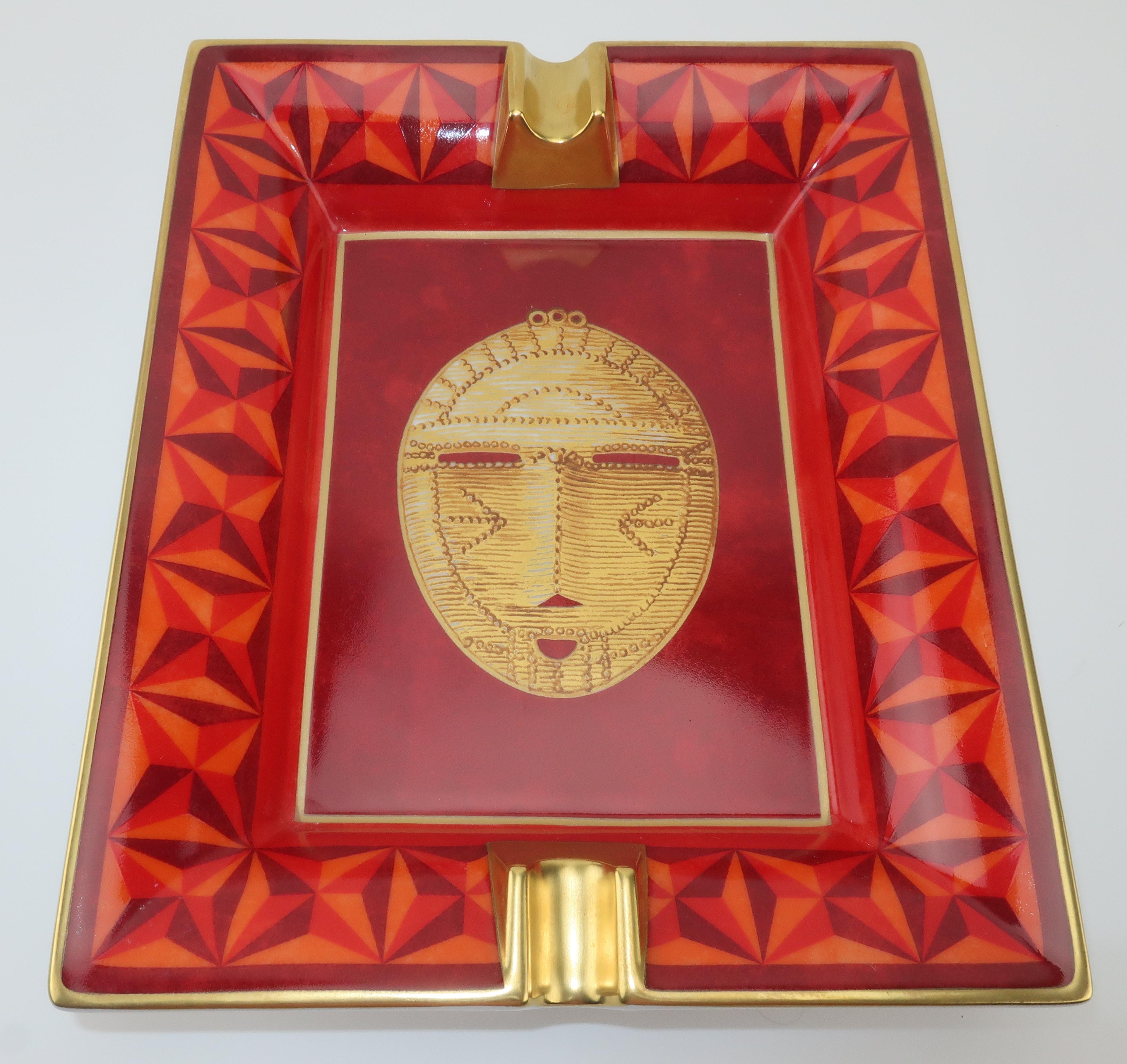 A vibrant and colorful Hermès porcelain ashtray in shades of red, orange and gold depicting a detailed hand painted mask with a geometric star decorated border.  Protected on the underside with a burgundy velvet goatskin base and hallmarked on one