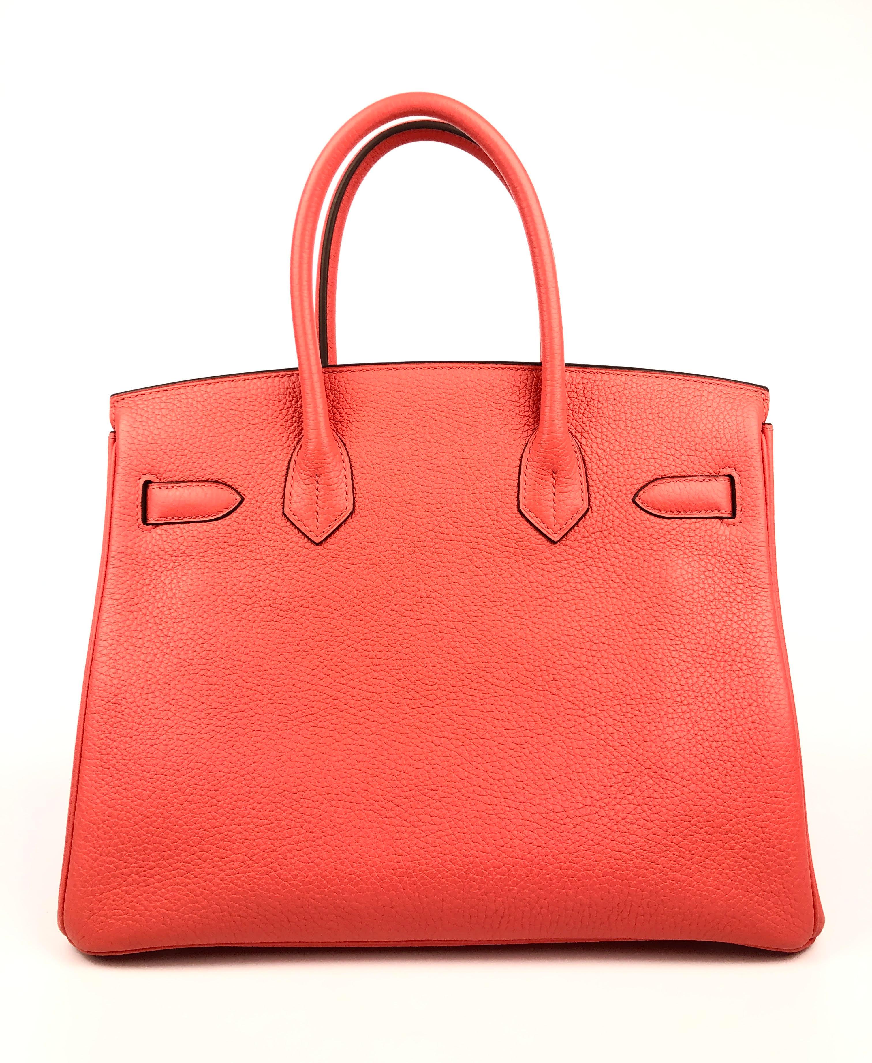 This authentic Hermès Punch Pink Red Togo 30 cm Birkin is in pristine unworn condition with the protective plastic intact on the hardware.  Waitlists exceeding a year are commonplace for the intensely coveted classic leather Birkin bag.  Each piece