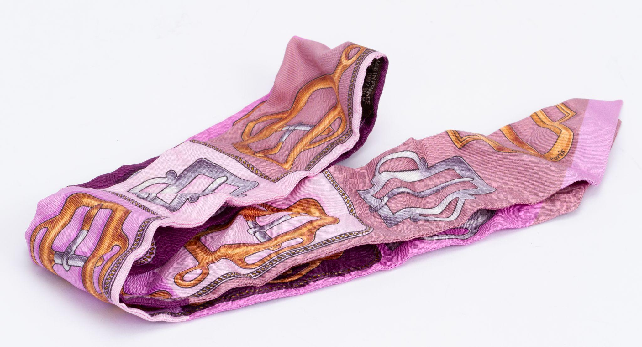New Hermès Trophy Ribbon Twilly. The scarf is made out of silk and the main color is pink. The print shows saddle stirrups. The pieces comes with a scarf ring. It is packed in the original box and ribbon.