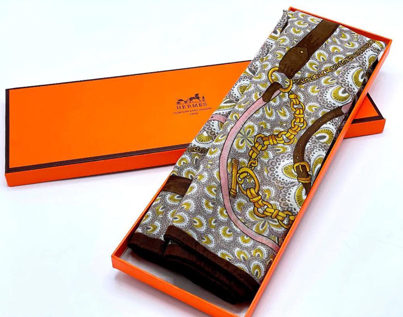 Foulard en soie Hermès dans sa boîte d'origine des années 1970, France

Cette écharpe présente un design équestre classique dans des tons marron, or, rose et gris, avec des détails de sangles et de chaînes sur fond de motif géométrique et floral. La