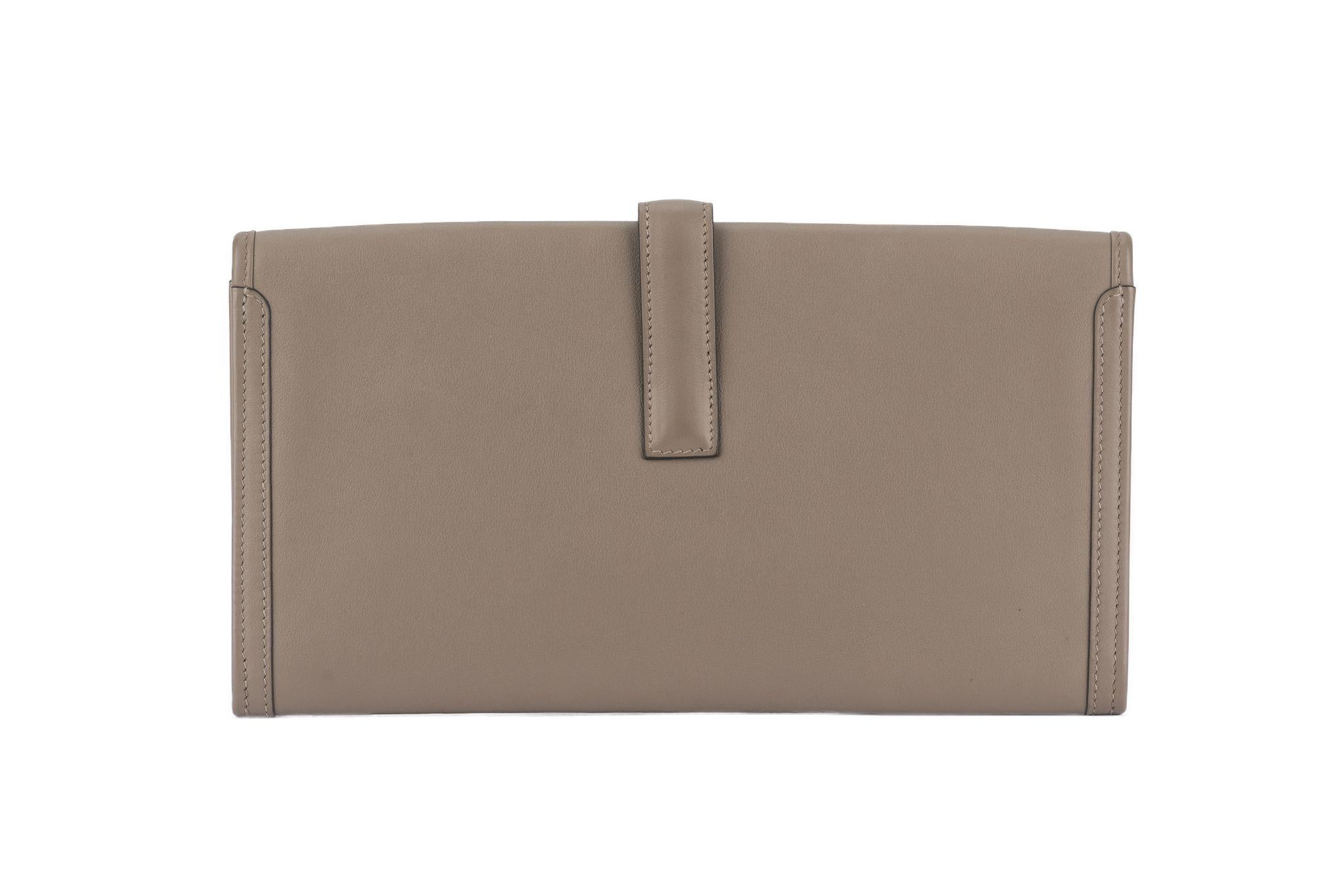 Questa pochette Hermès Swift Jige Elan 29 in Etoupe è realizzata in pelle di vitello a grana morbida di colore grigio. La patta e il cinturino incrociati si fissano sotto una 