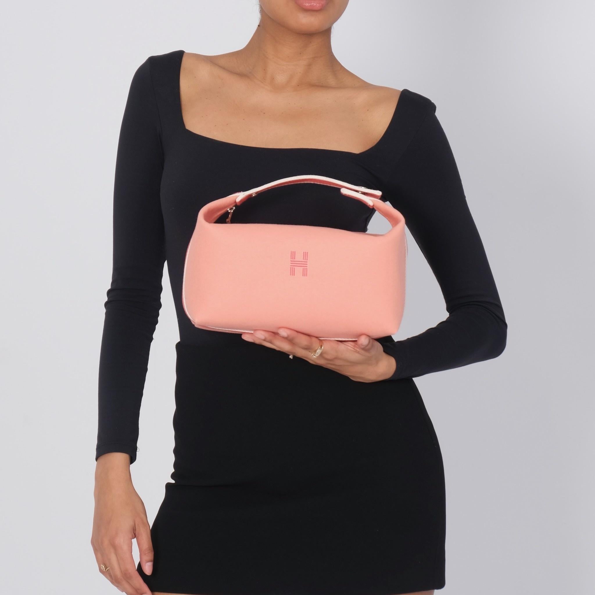 This vanity pouch bag is constructed of pale orange (supplier color name is Goyave) canvas with a contrasting dark orange & white stripe around the bag. The bag features a top handle strap with snap closure, top zipper closure and a front logo. The