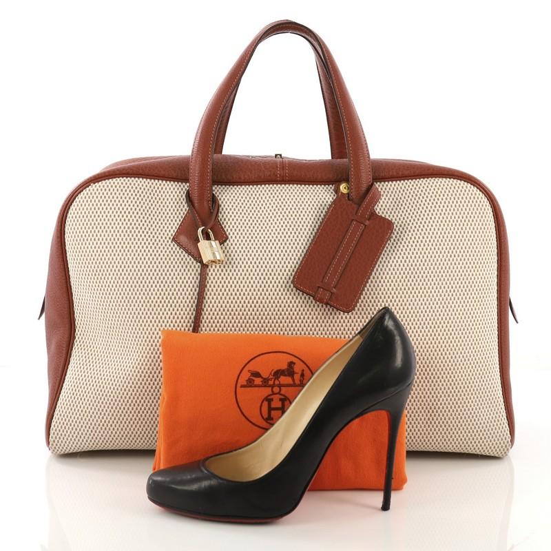 This Hermes Victoria II Handbag Toile 43, crafted in Marron Glace brown Buffalo Skipper & Ecru beige Toile GM, features dual top leather handles, protective base studs, and gold hardware. Its zip closure opens to a beige canvas interior with a zip