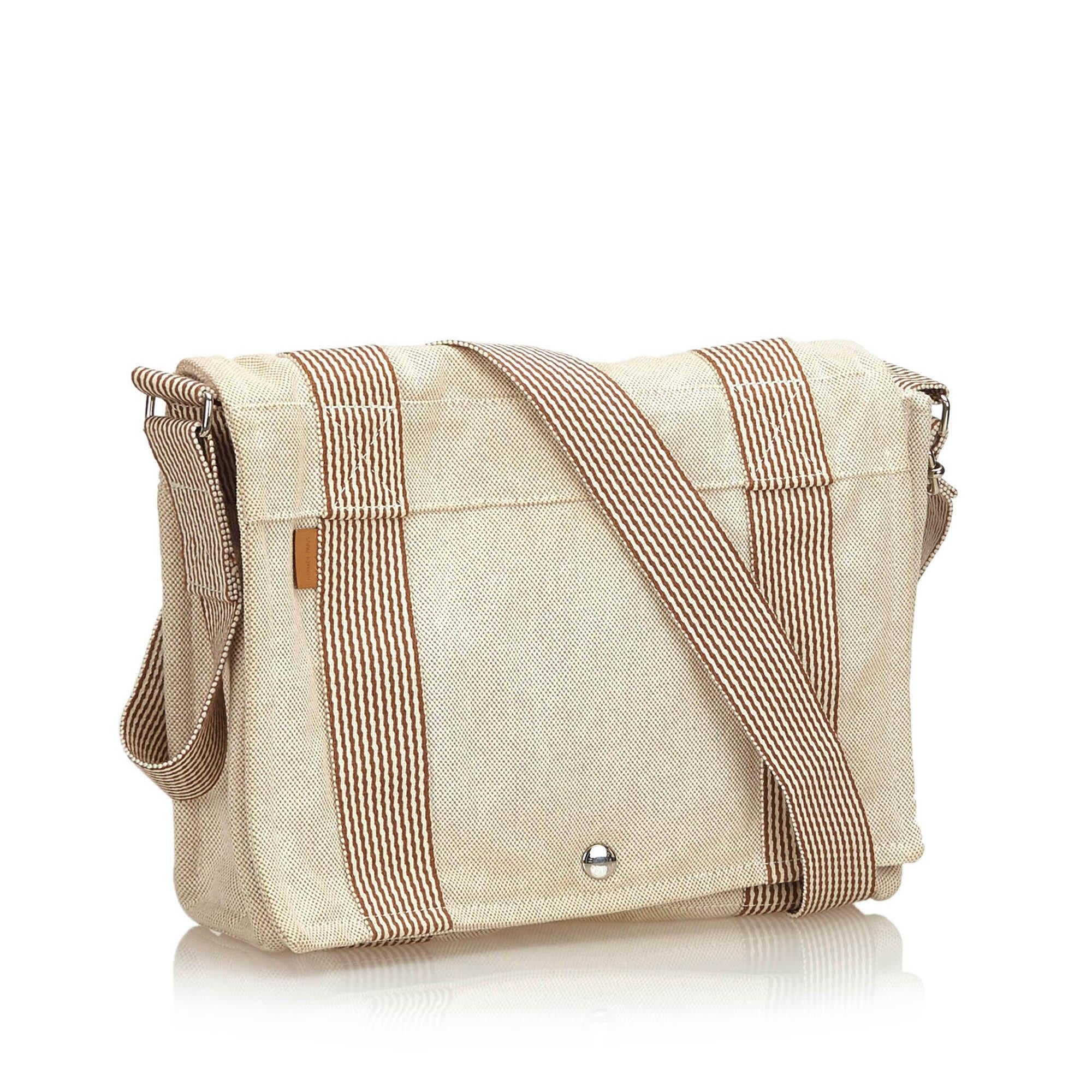 This shoulder bag features a canvas body, flat strap, front flap with button closure and interior zip pocket. It carries as B condition rating.

Inclusions: 
Box

Dimensions:
Length: 23.50 cm
Width: 30.00 cm
Depth: 8.00 cm
Shoulder Drop: 51.50