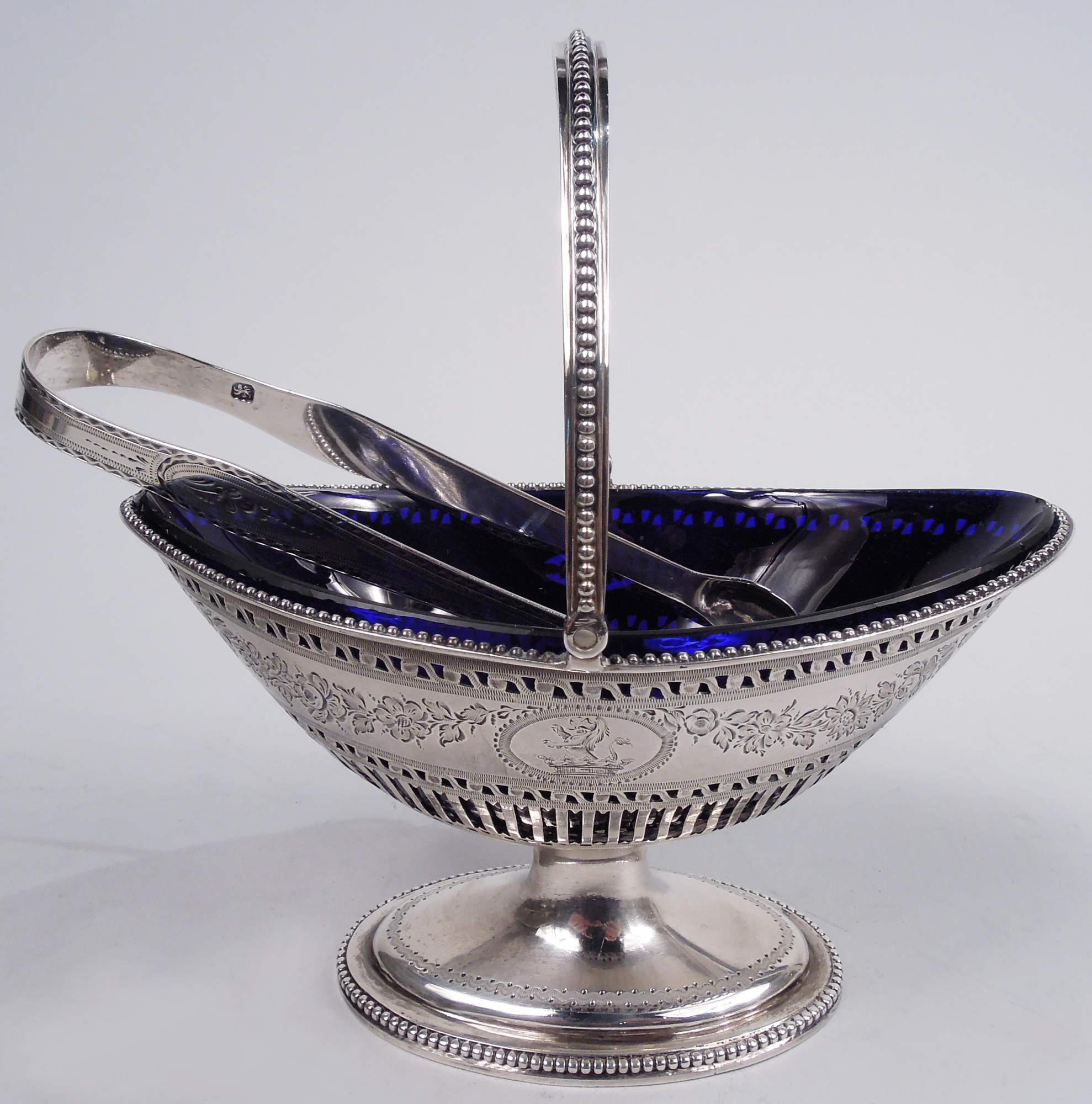 George III sterling silver sugar basket. Made by Hester Bateman in London in 1784. Ovoid bowl with c-scroll swing handle and raised foot. Pierced colonnade and rinceaux as well as ovals inset with paterae. Engraved garland. Beading. Cobalt glass