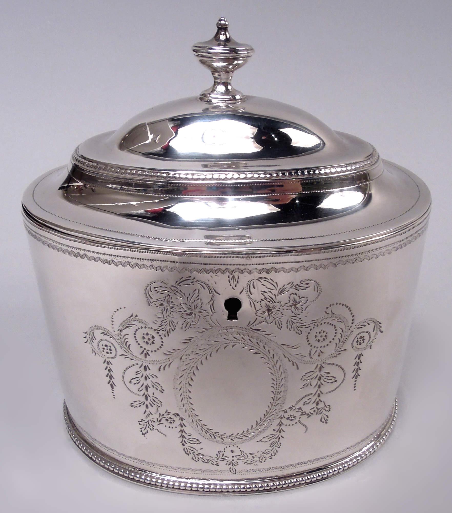 George III sterling silver tea caddy with spoon. Made by Hester Bateman in London in 1784. Oval bowl with straight sides. Cover hinged and domed with vasiform finial. On front, bright-cut oval wreath (vacant) surrounded by leafing scrollwork,