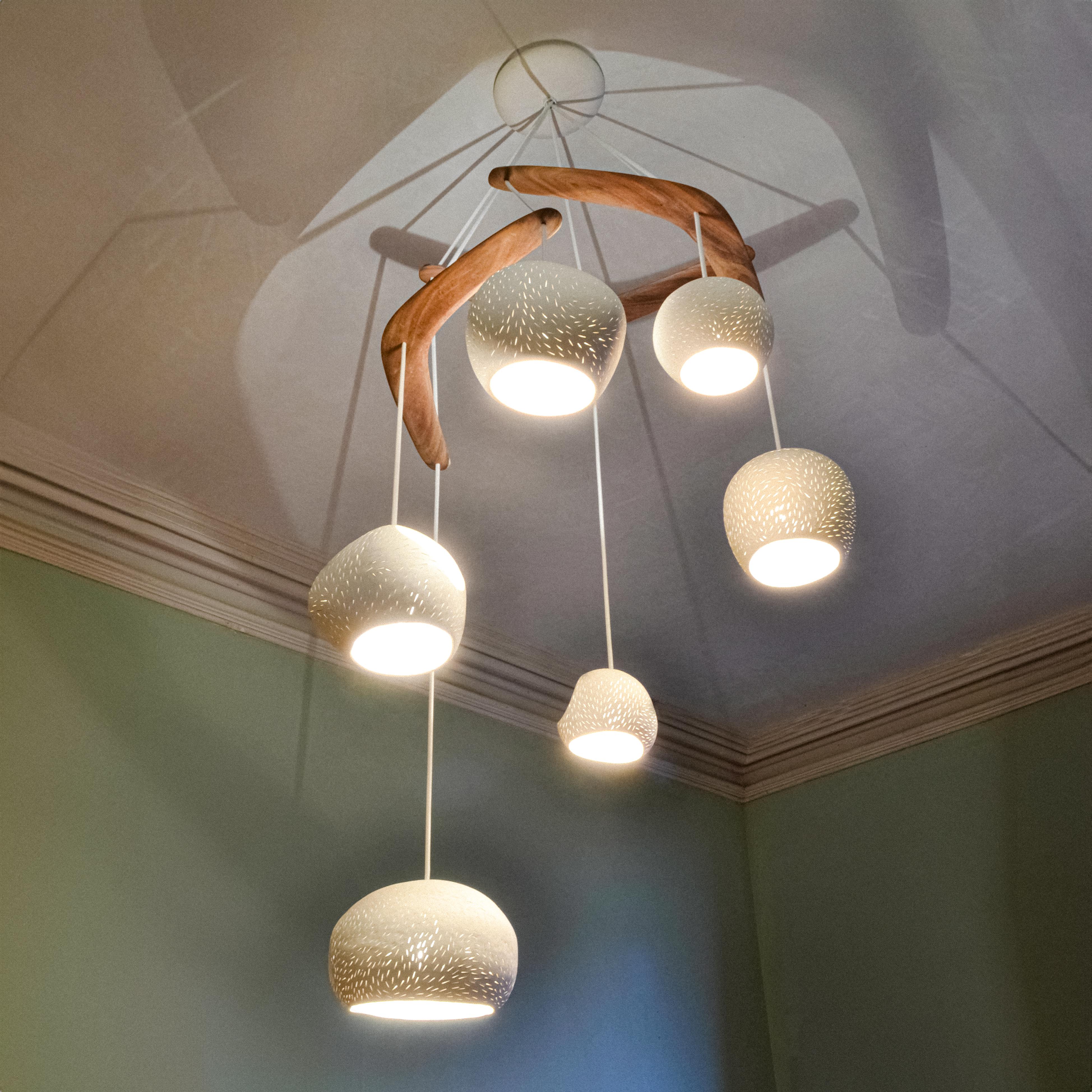 Ce lustre moderne est un éclairage sculptural en céramique. Le design fonctionnel comprend six lampes suspendues en céramique blanc chaud mat qui sont suspendues à du bois brigalow australien courbé, qui ensemble créent un mobile sculptural de