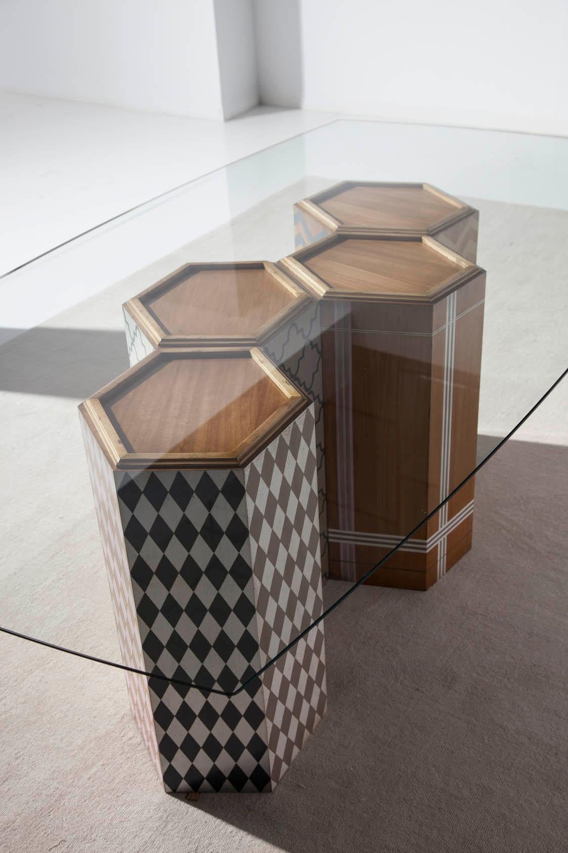 Table base made up of four hexagonal columns, decorated with different motifs and colours. The glass envelope, which is manufactured in any size, takes a slightly curved shape on all four sides. Architectural and light effect that does not