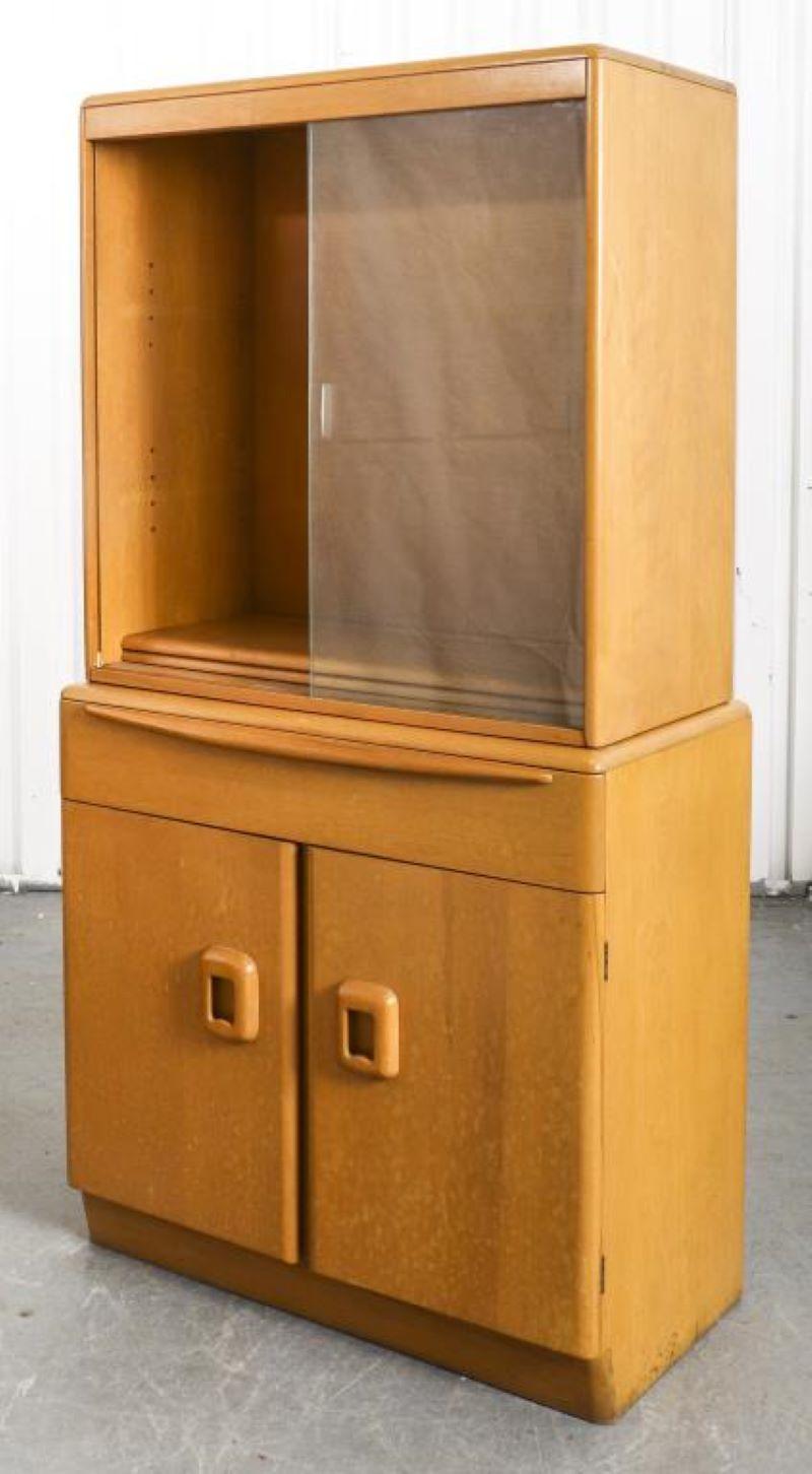 Mid-century Modern blonde wood display cabinet by Heywood Wakefield, with glass doors and two shelves atop drawers, signed to reverse. 64” H x 32” W x 17” D.