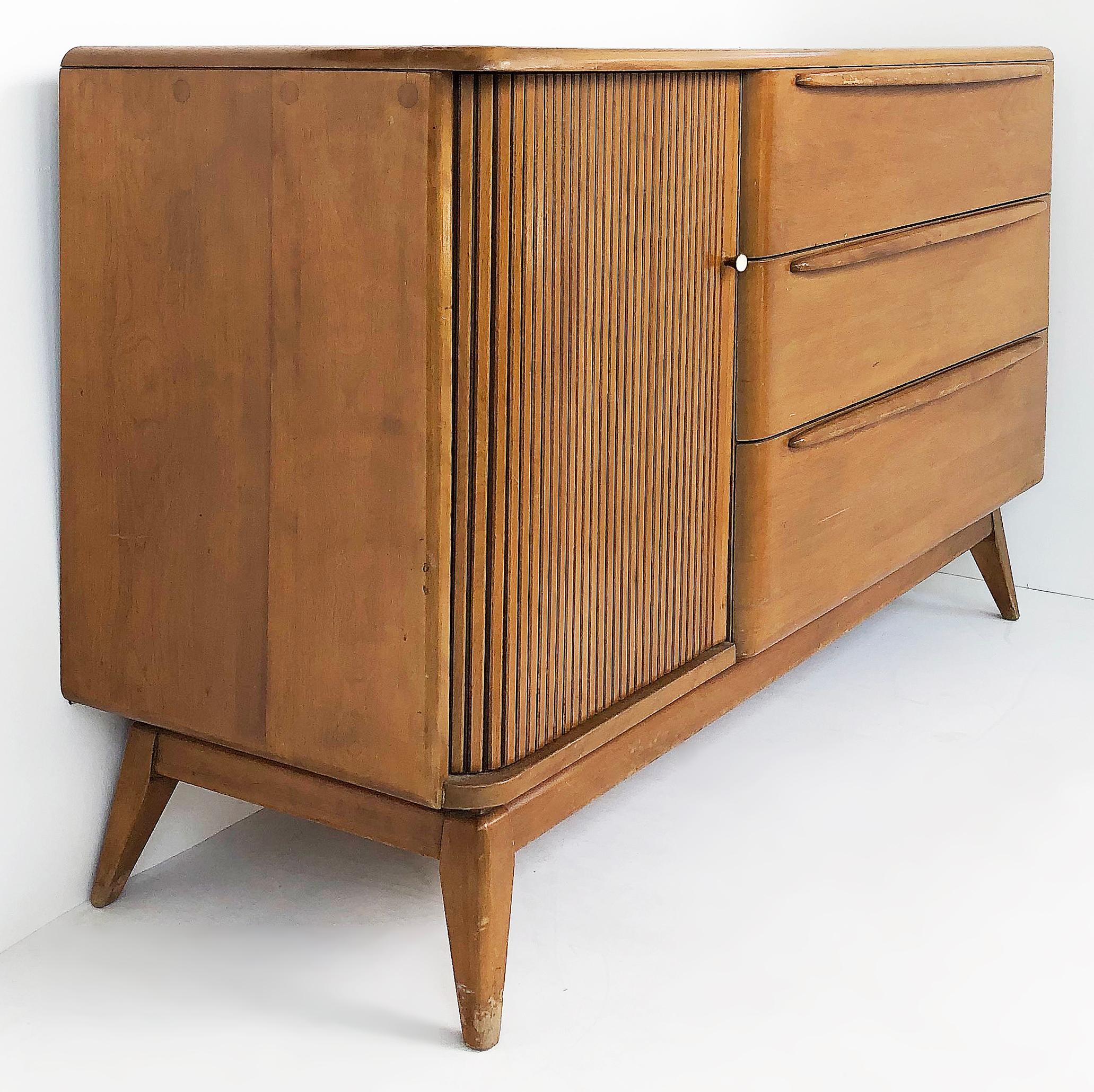 Offered for sale is a Heywood-Wakefield credenza cabinet with a tambour door that opens to reveal open shelves and three drawers to the right.
