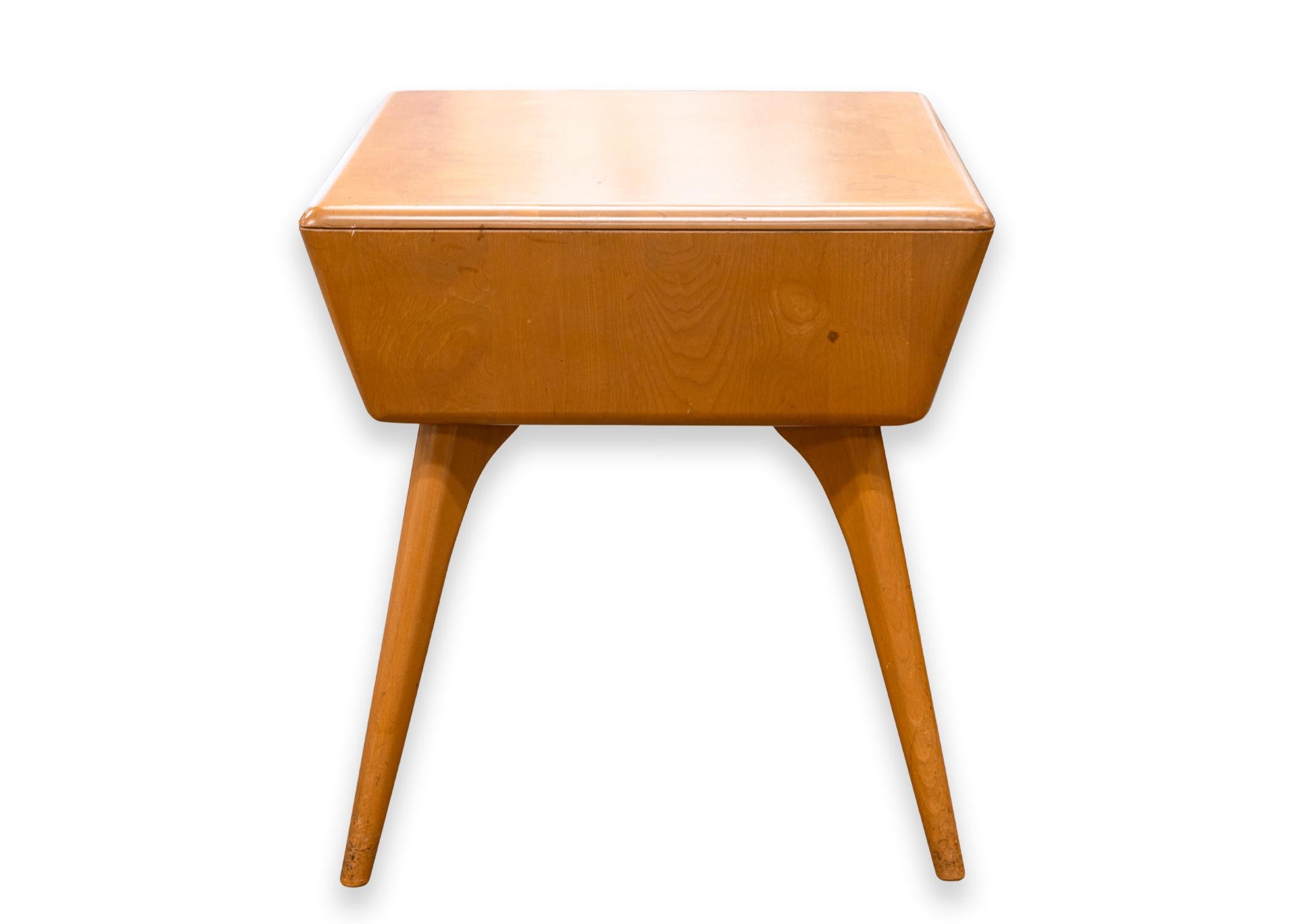 A Heywood Wakefield Encore side end table. A lovely mid century piece of furniture featuring a solid wood construction with a Champagne wood finish, rounded legs, and a versatile open storage compartment. This table is stamped on the underside with
