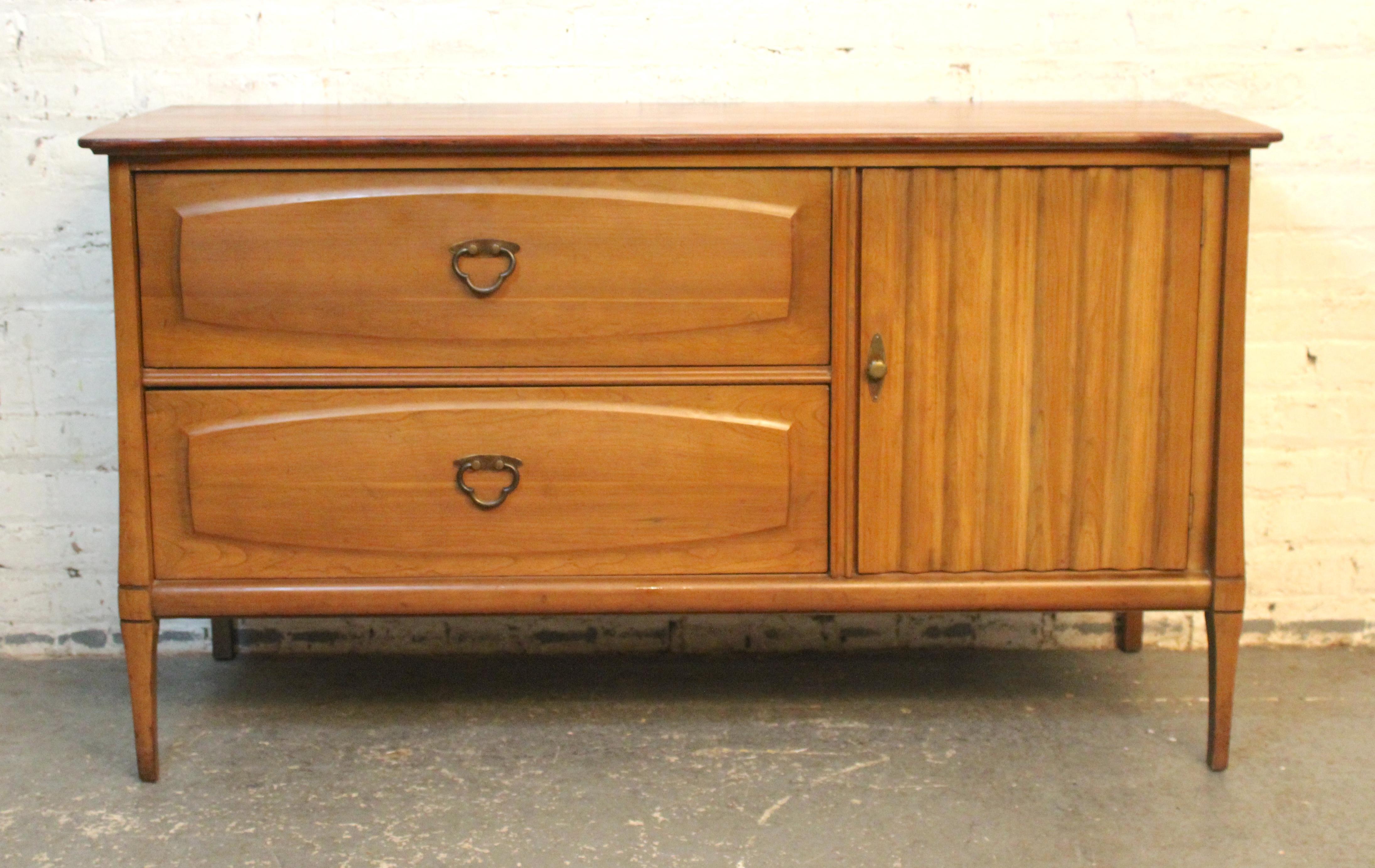 This mid-century modern sideboard features a smaller footprint than most, but offers ample storage. Made of blonde maple wood and accenting brass clover shaped hardware, with two wide drawers and a single side cabinet. The simple design is adorned