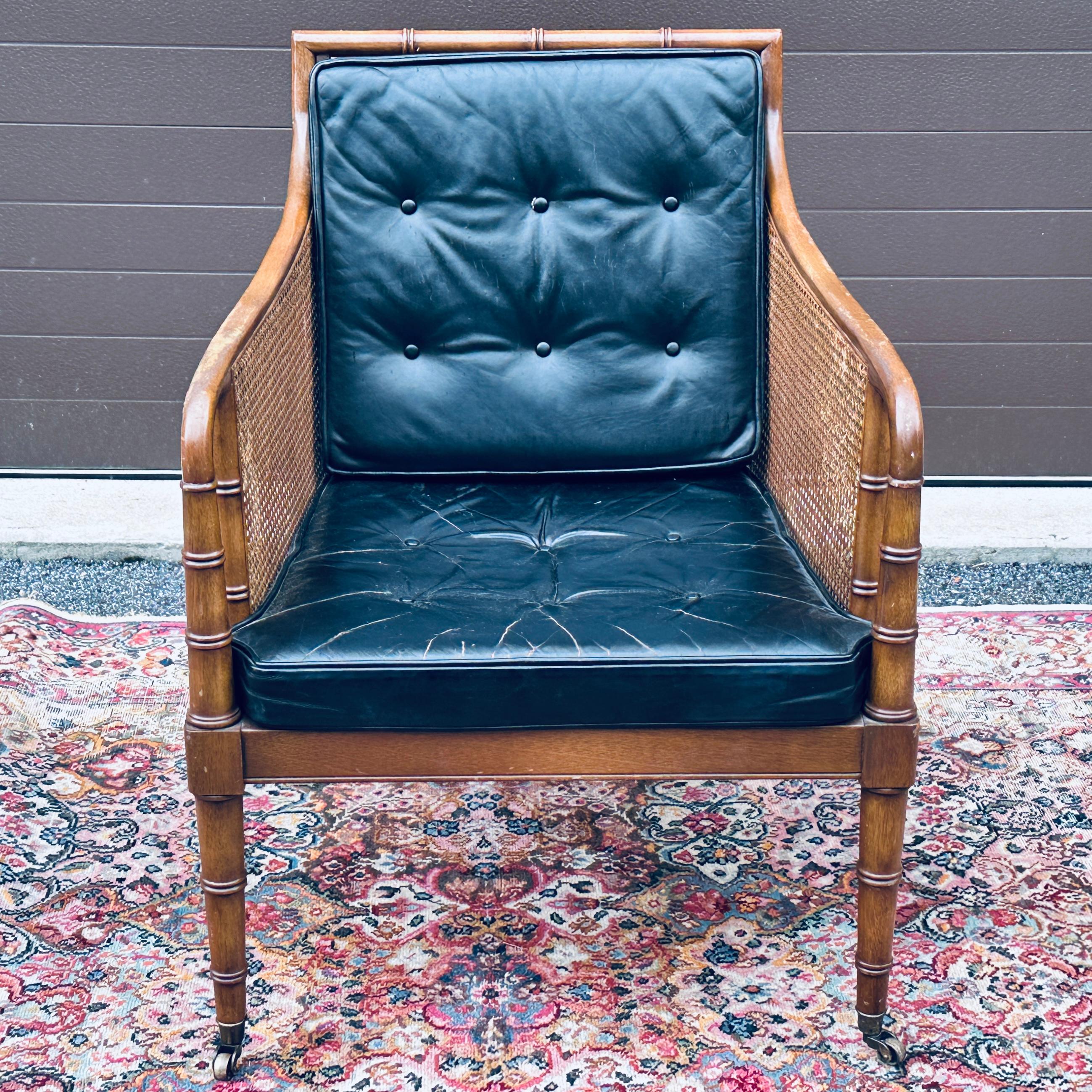 Classic Hickory Chair Regency style lounge chair featuring a carved hardwood faux bamboo frame with caned back and sides and black leather cushions. Front legs on brass casters. 

From smoke free estate with wear from age and use. Frame solid and