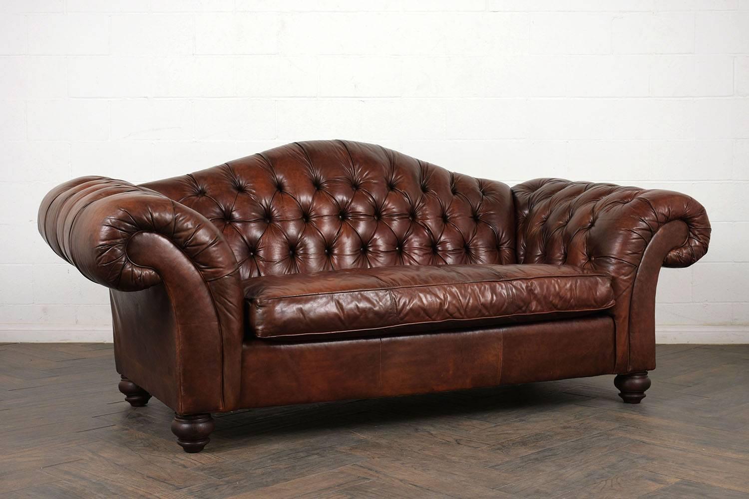 This 1970s Chesterfield sofa has a camel back profile with exaggerated scroll arms. The sofa is upholstered in a rich brown color with tufted details on the back and arms with single piping trim details. There is a single comfortable cushion that