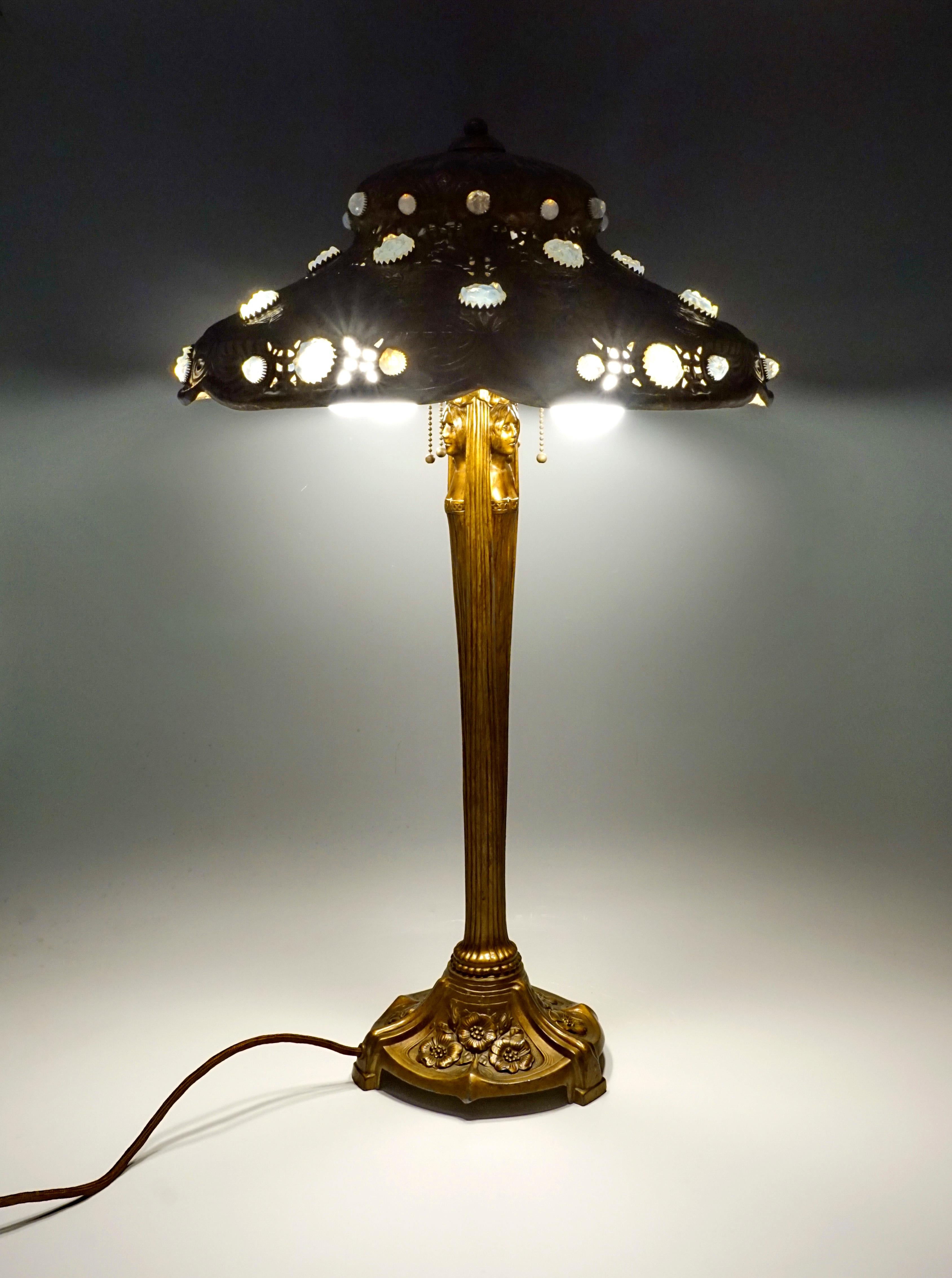 High table lamp on a round base divided into three segments filled with floral decorations, slender, high, widening, profiled column with embedded caryatids, hat-shaped, stepped metal shade with embossed floral elements and leaves, as well as