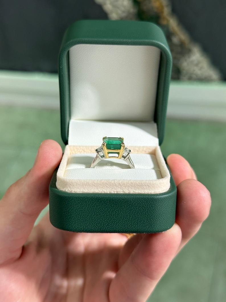 Introducing our stunning emerald and diamond three-stone ring, featuring a captivating 4.49-carat natural asscher cut emerald sourced from Zambia, renowned for its exceptional color, clarity, and luster. Set in an elegant four double-claw prong