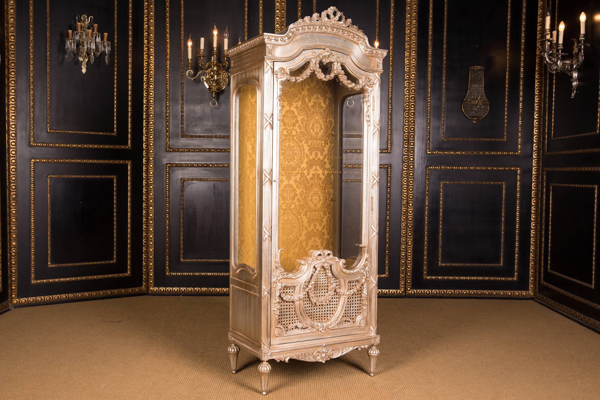 Massive beech, poliment silver. High-angled, three-sided, facet-cut glazed, one door body on conical legs. The door is partially covered with wickerwork. The front, corners and sides are decorated with carved, classical elements such as garlands and