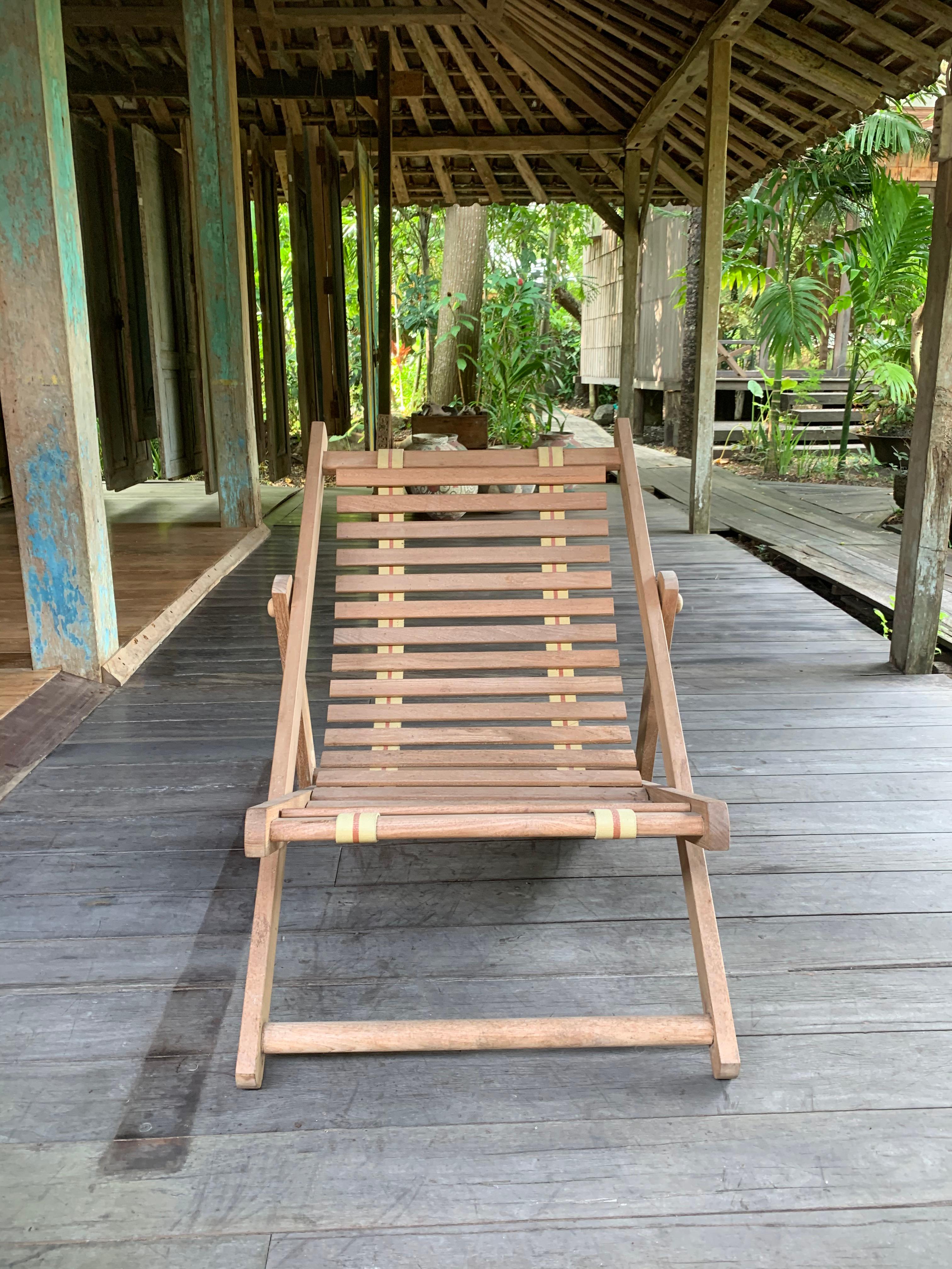 A hand-crafted, foldable teak lounger chair. These chairs are crafted by local artisans and features a subtle, smooth finished wood texture. The straps which hold the horizontal wooden strats of the seat are secured by two reclaimed motor belts from