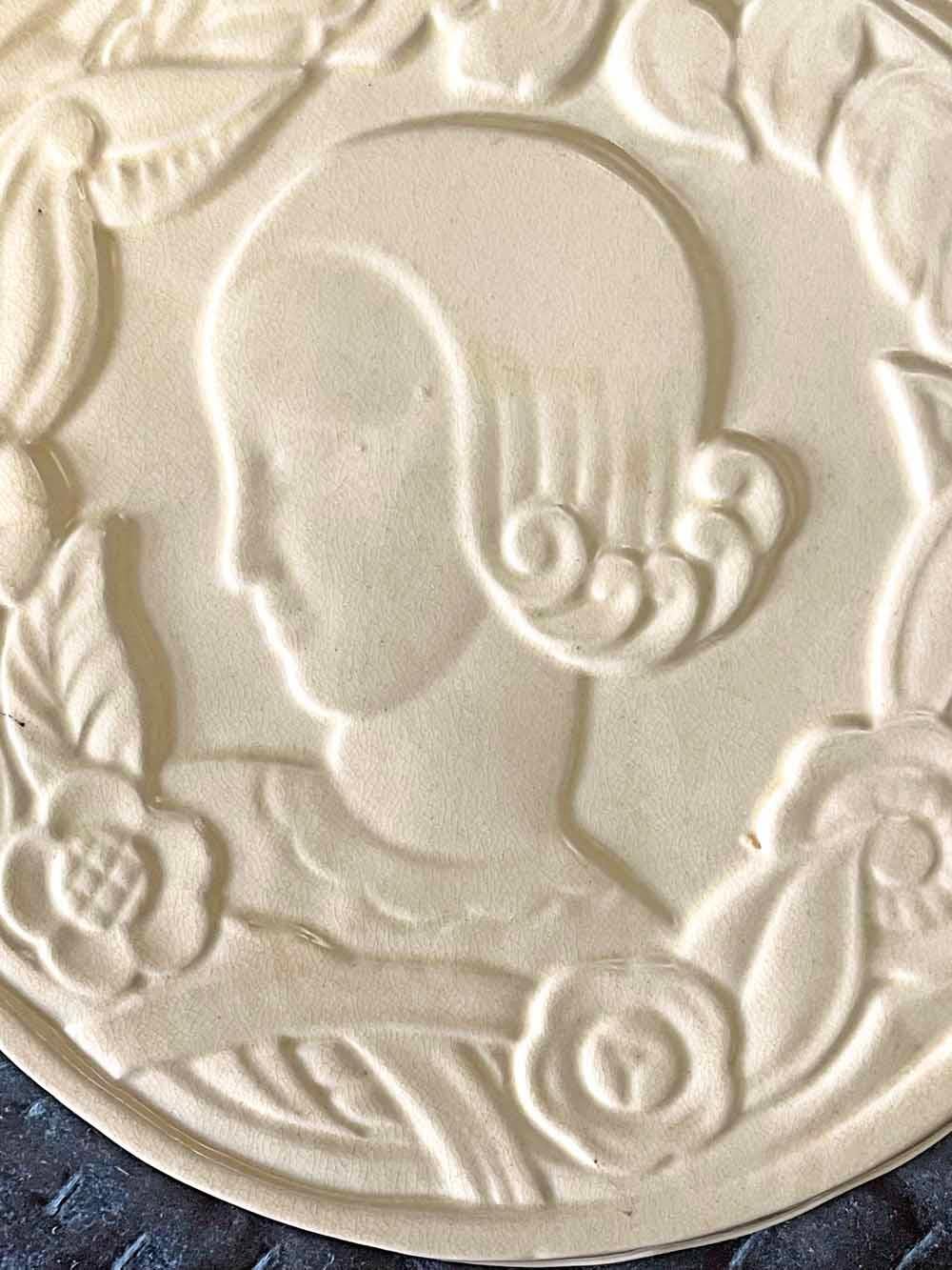 Beautifully enrobed in a pale, ivory-toned glaze, this rare, high style Art Deco trivet depicts a fashionable female figure in profile, her hair curled in repeating waves like the lapping water of an seaside beach.  The woman is surrounded by a